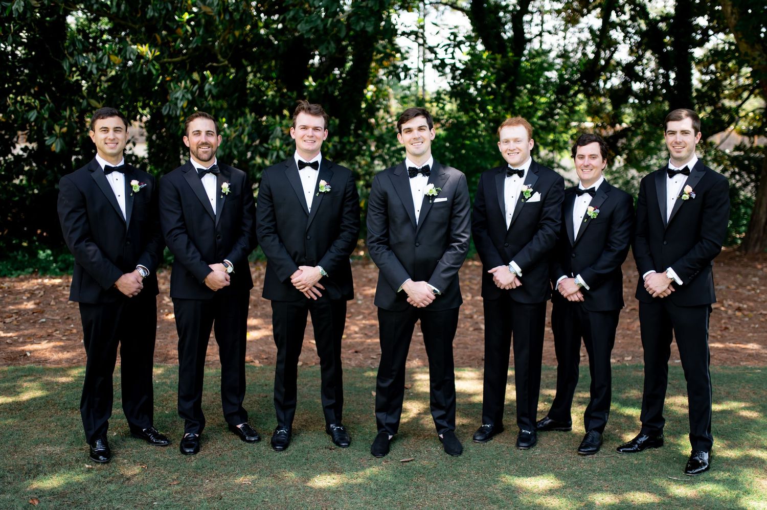 Seven groomsmen in matching black tuxedos pose outdoors under lush green trees.