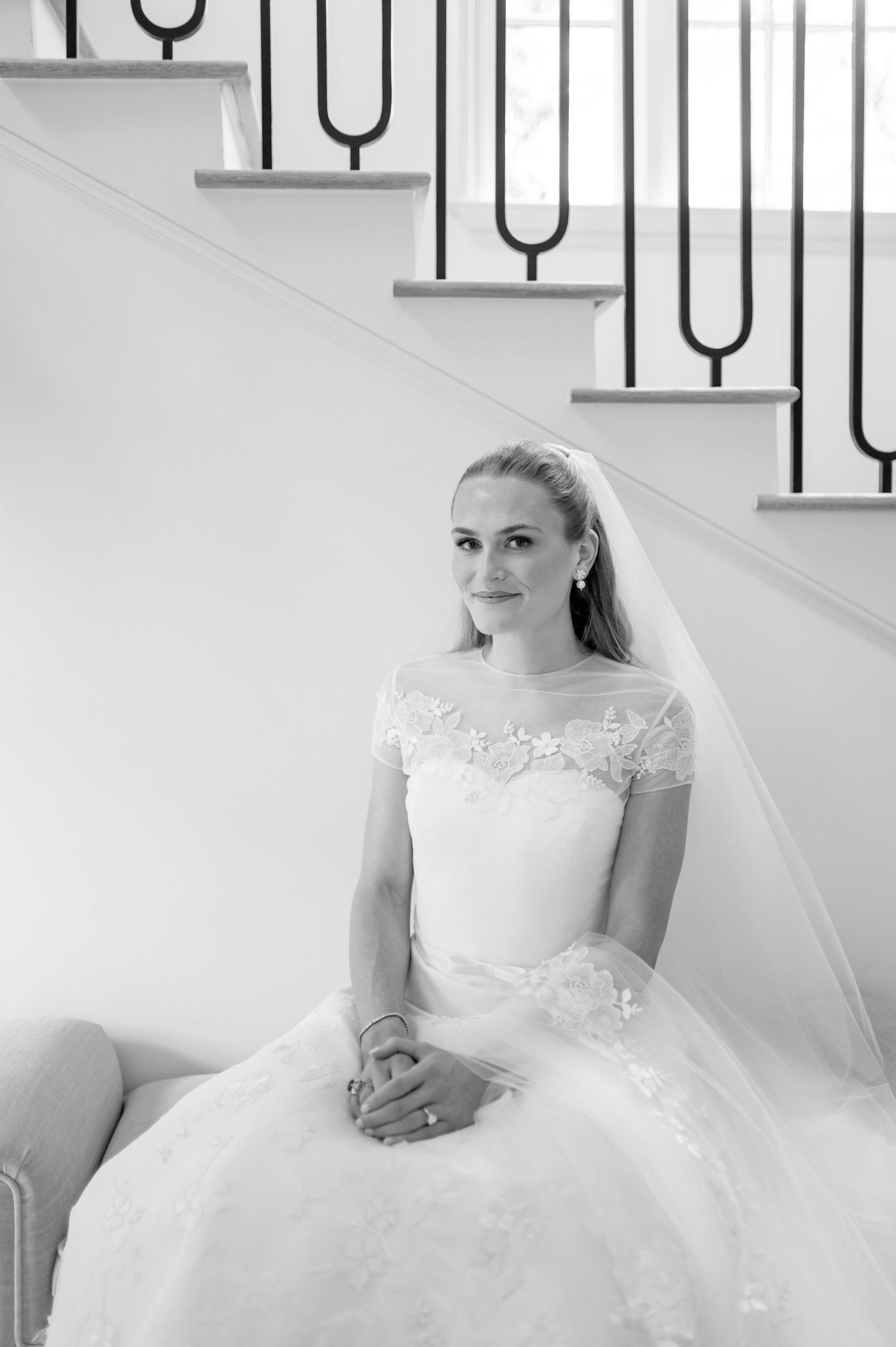 Elegant bride in lace off-shoulder wedding gown and veil seated on stairs in black and white portrait.