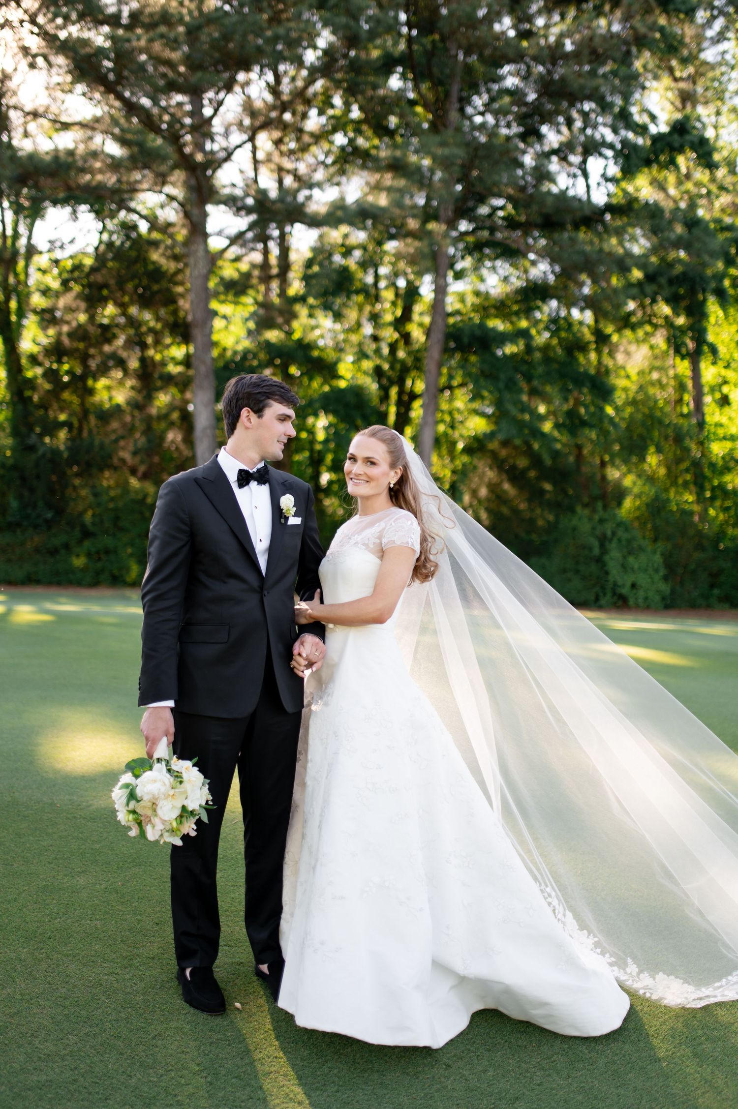 Bride and groom portrait outdoors with flowing veil and white bouquet on golf course.