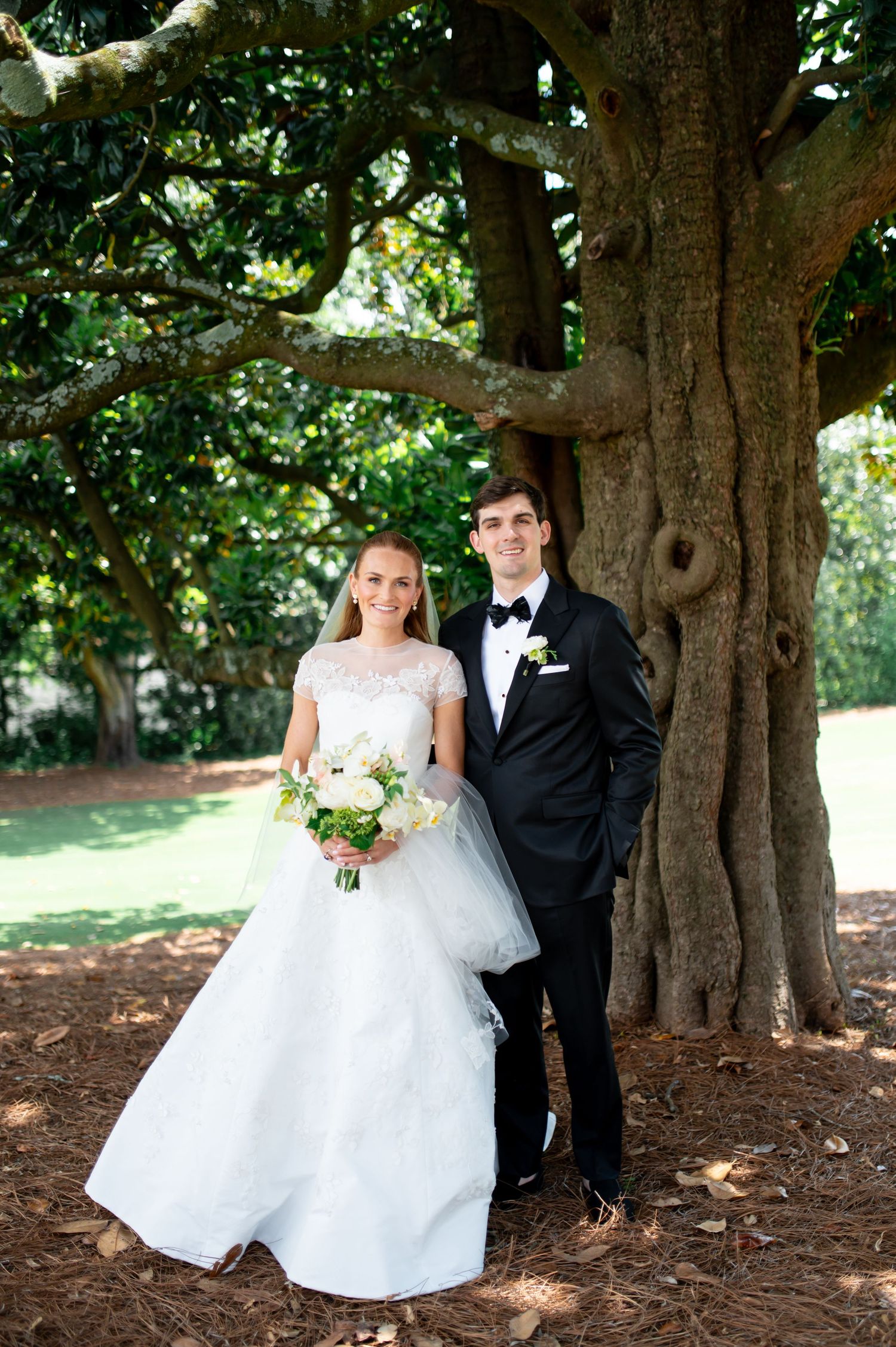 Bride and groom portrait under large oak tree, bride holding white bouquet, groom in black tuxedo.