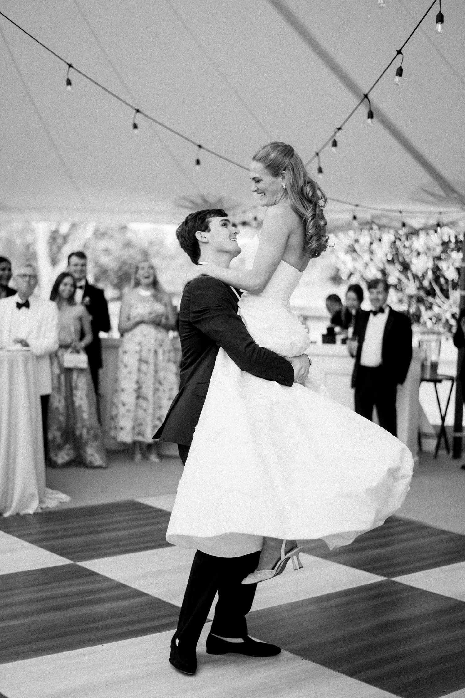 Groom lifts bride joyfully during first dance under tent with string lights in black and white.