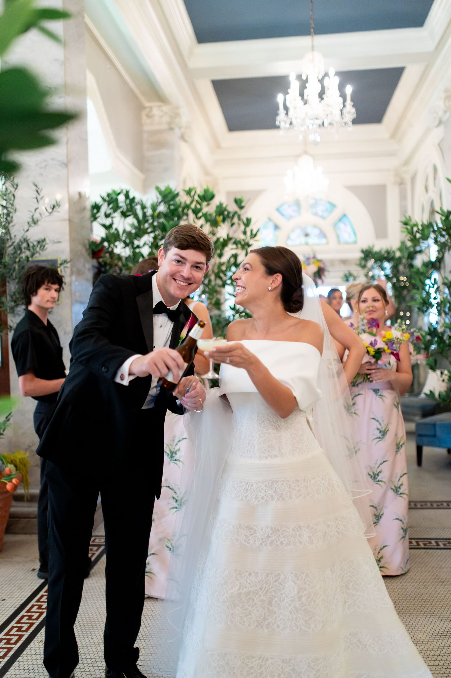 Joyful newlyweds toasting champagne in ornate venue lobby adorned with greenery and chandelier.