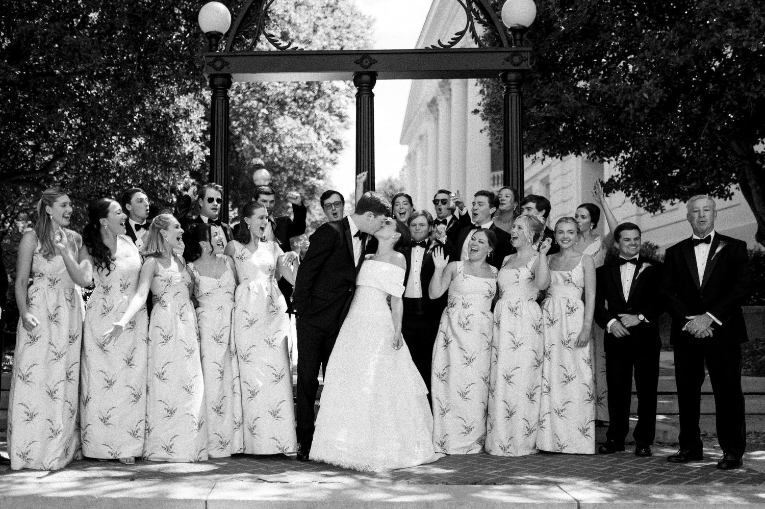 Black and white wedding photo of bride and groom kissing surrounded by large bridal party in floral dresses and tuxedos.