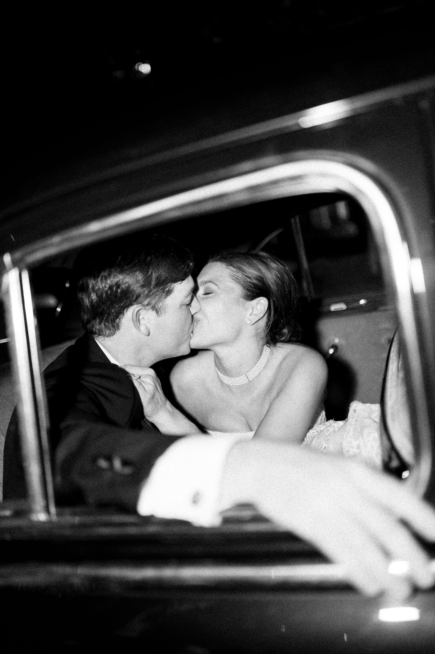 Bride and groom share a romantic kiss inside vintage car in black and white wedding portrait.