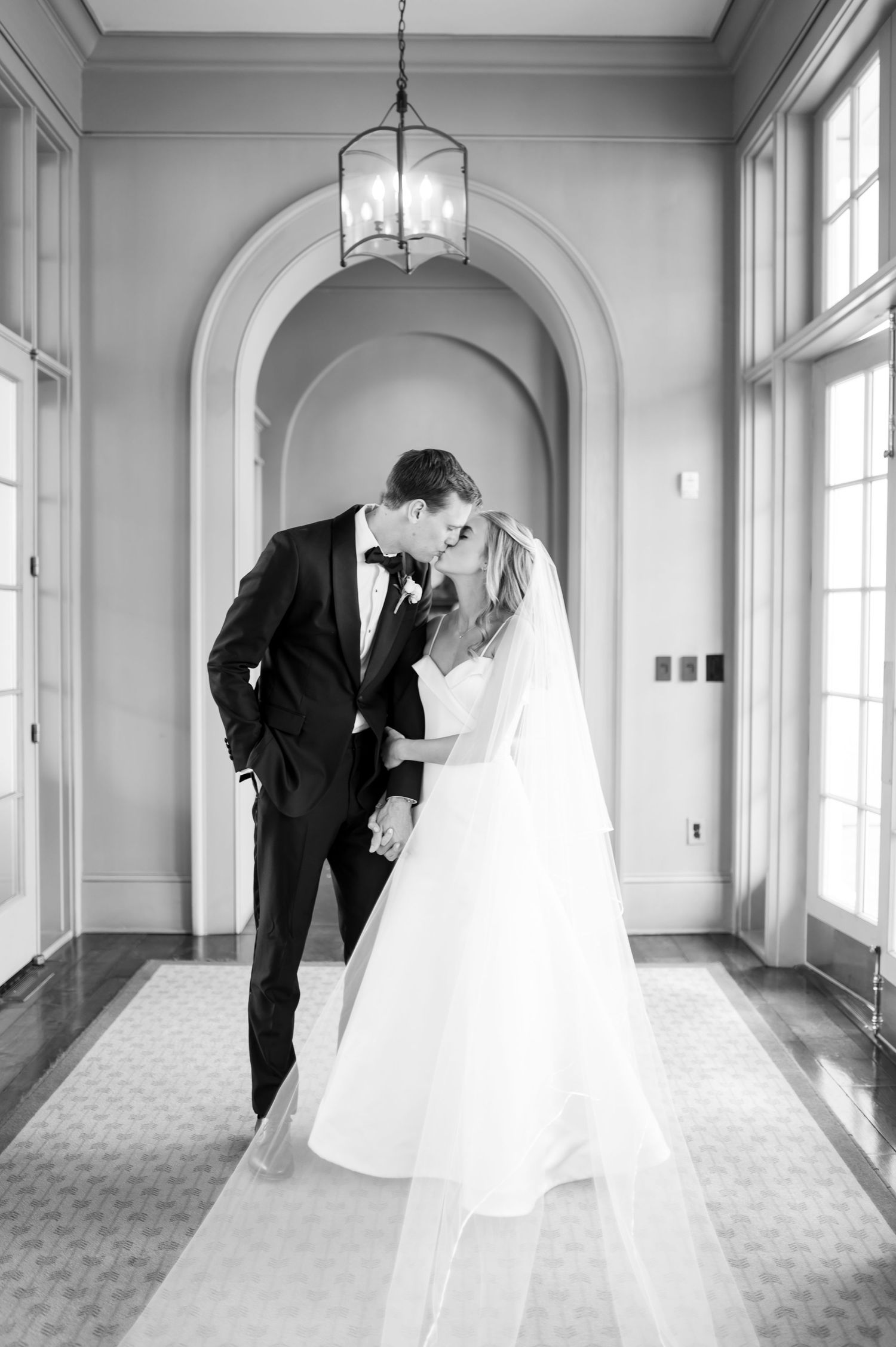 Elegant wedding couple shares intimate moments in a bright hallway with arched windows and classic architecture.
