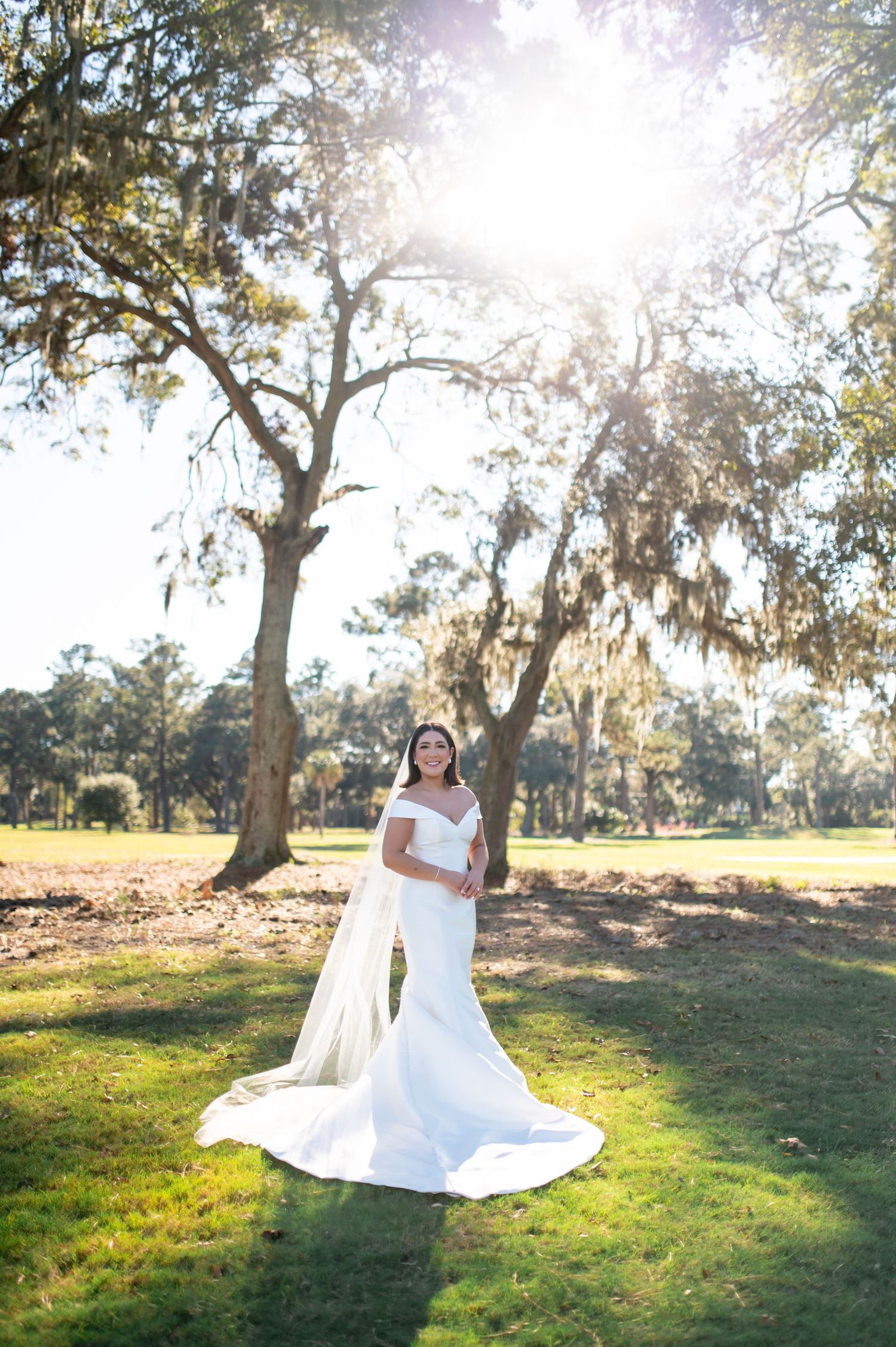 Bride in elegant white gown posing in sunlit field with trees.