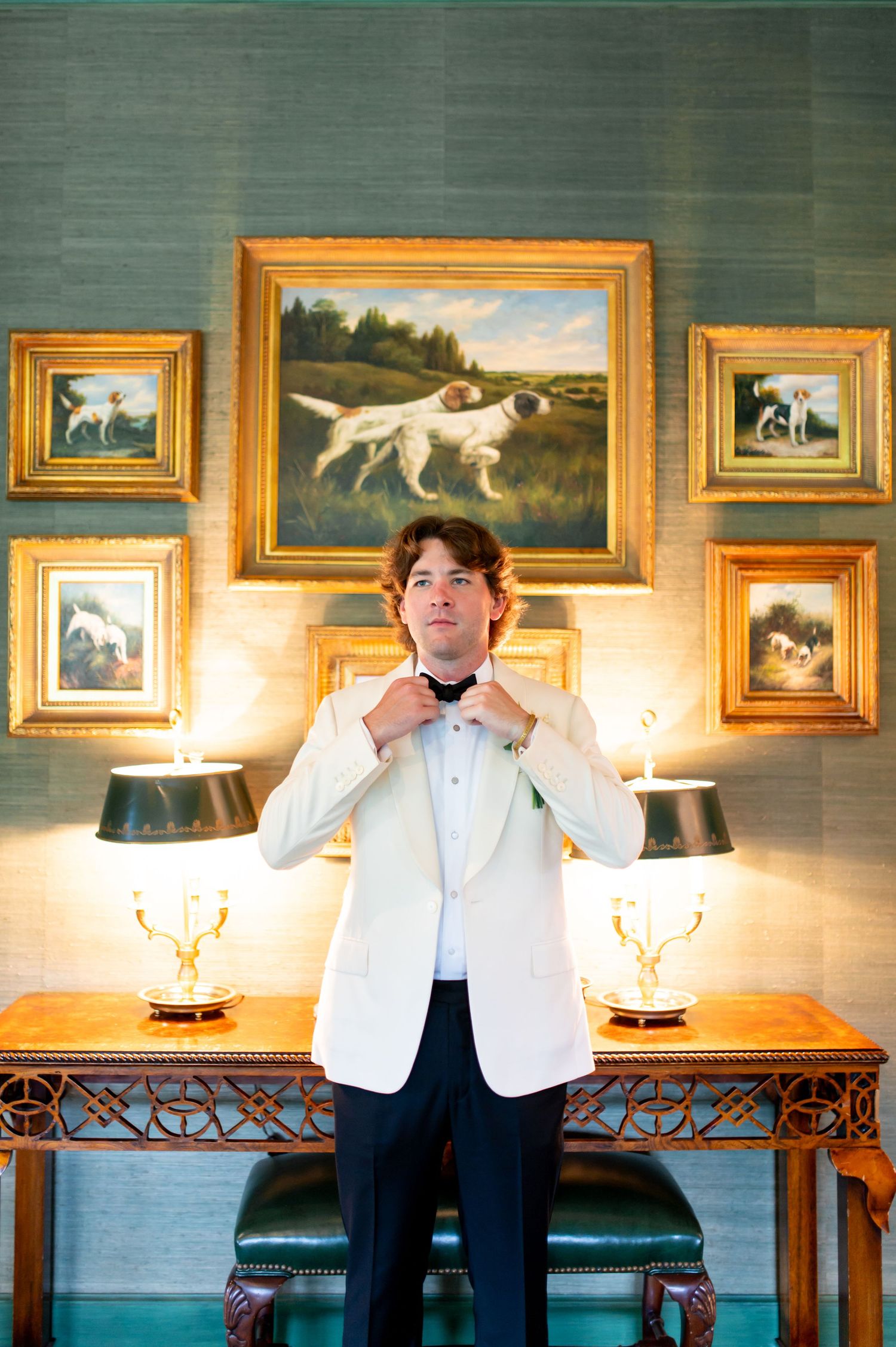 Person in white formal attire adjusts bow tie in room with hunting paintings and green wallpaper.
