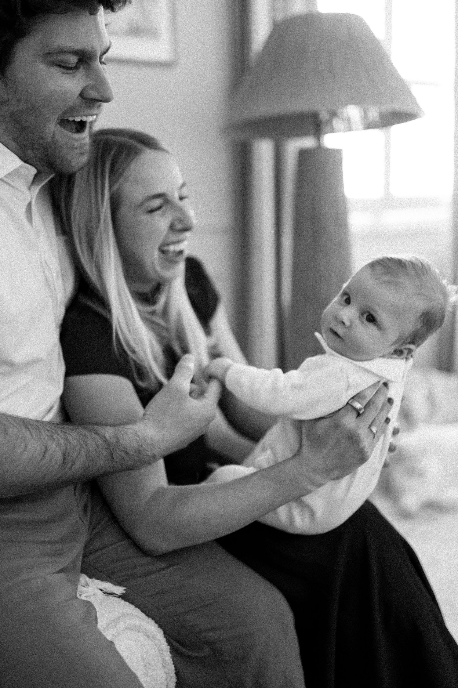 Black and white photo of a joyful family moment with a baby and adults laughing together in a cozy home setting.