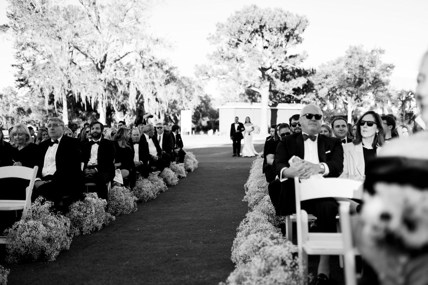 Black and white photo of an outdoor wedding ceremony with rows of white chairs and floral decorations.