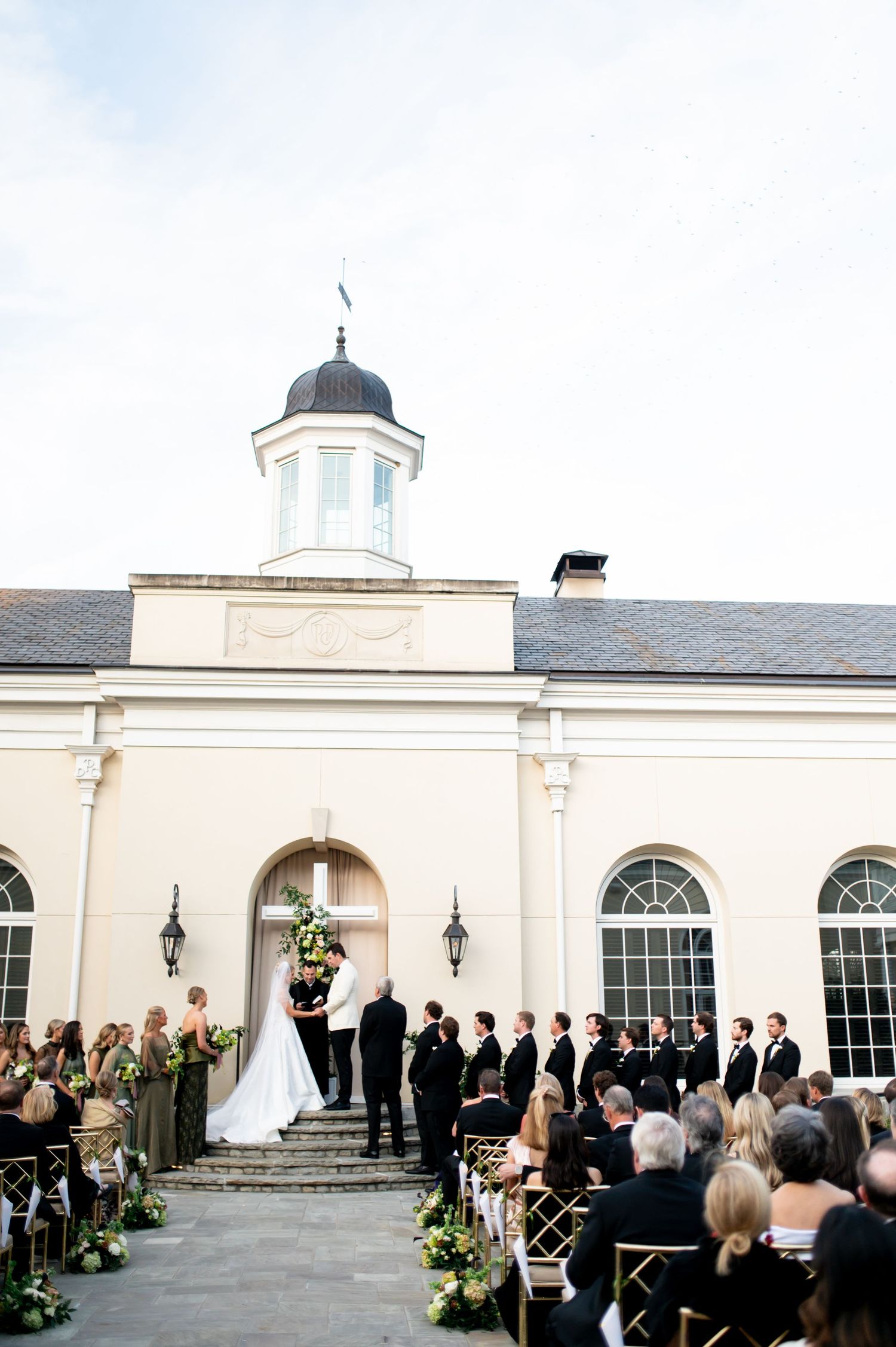 Outdoor wedding ceremony in progress at elegant colonial-style venue with white columns.