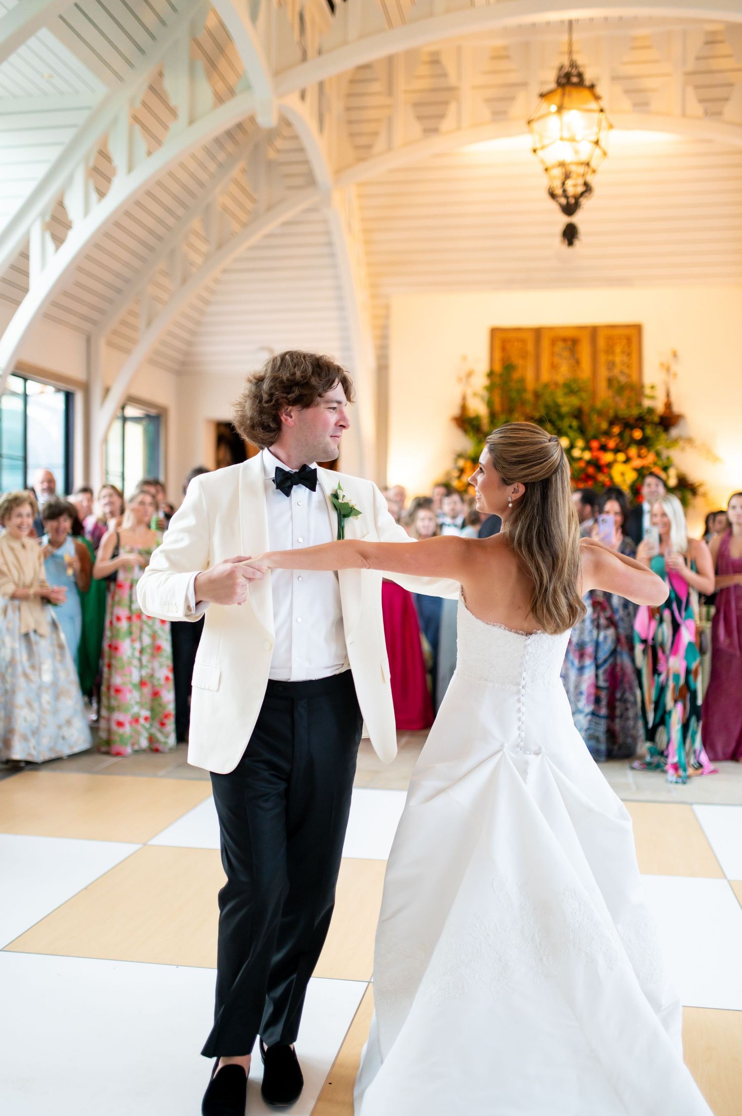A couple shares their first dance at a wedding reception in an elegant white-beamed venue with a chandelier overhead.