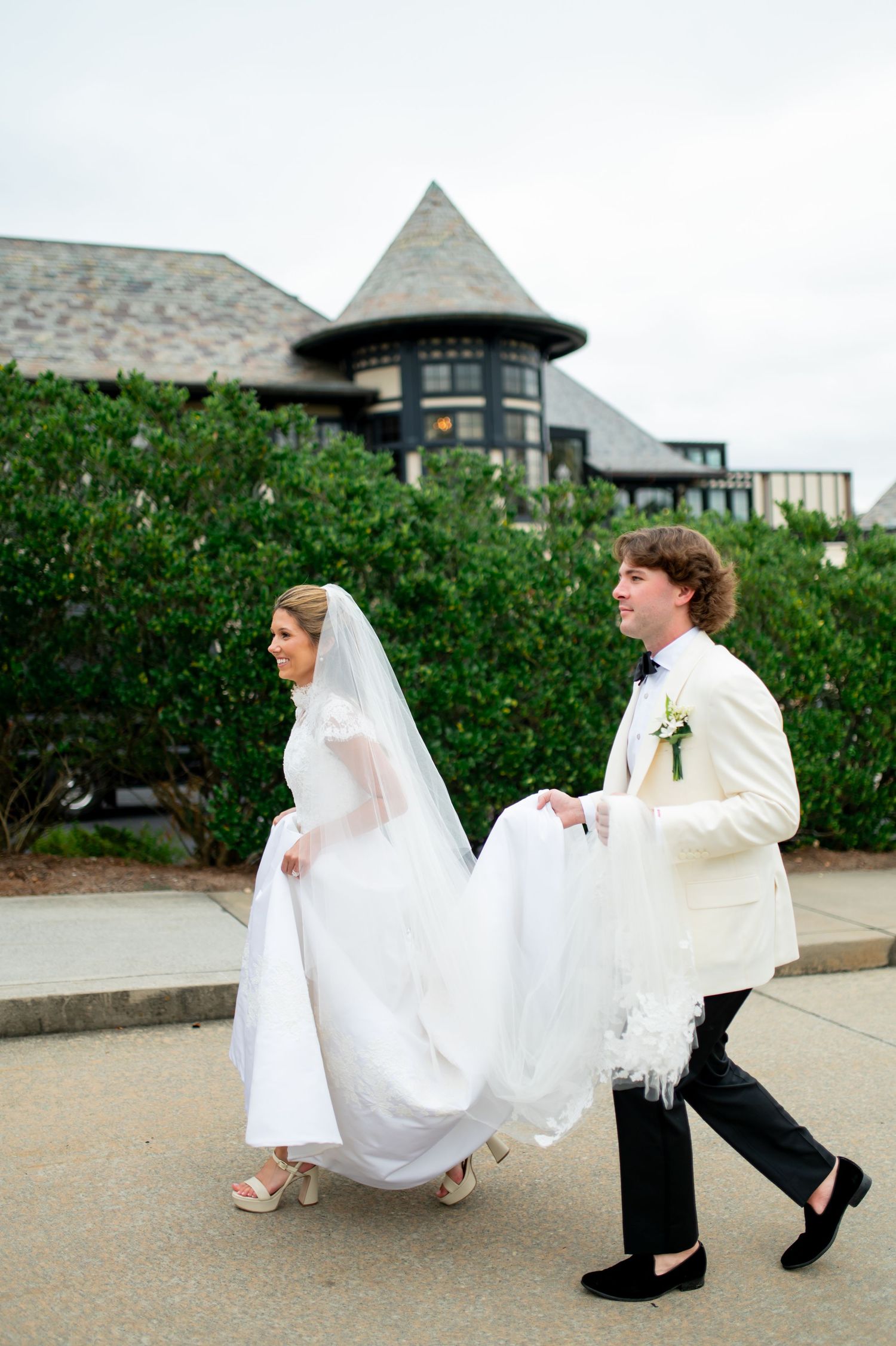 Wedding couple walking outside historic stone building with groom helping carry bride's dress train.
