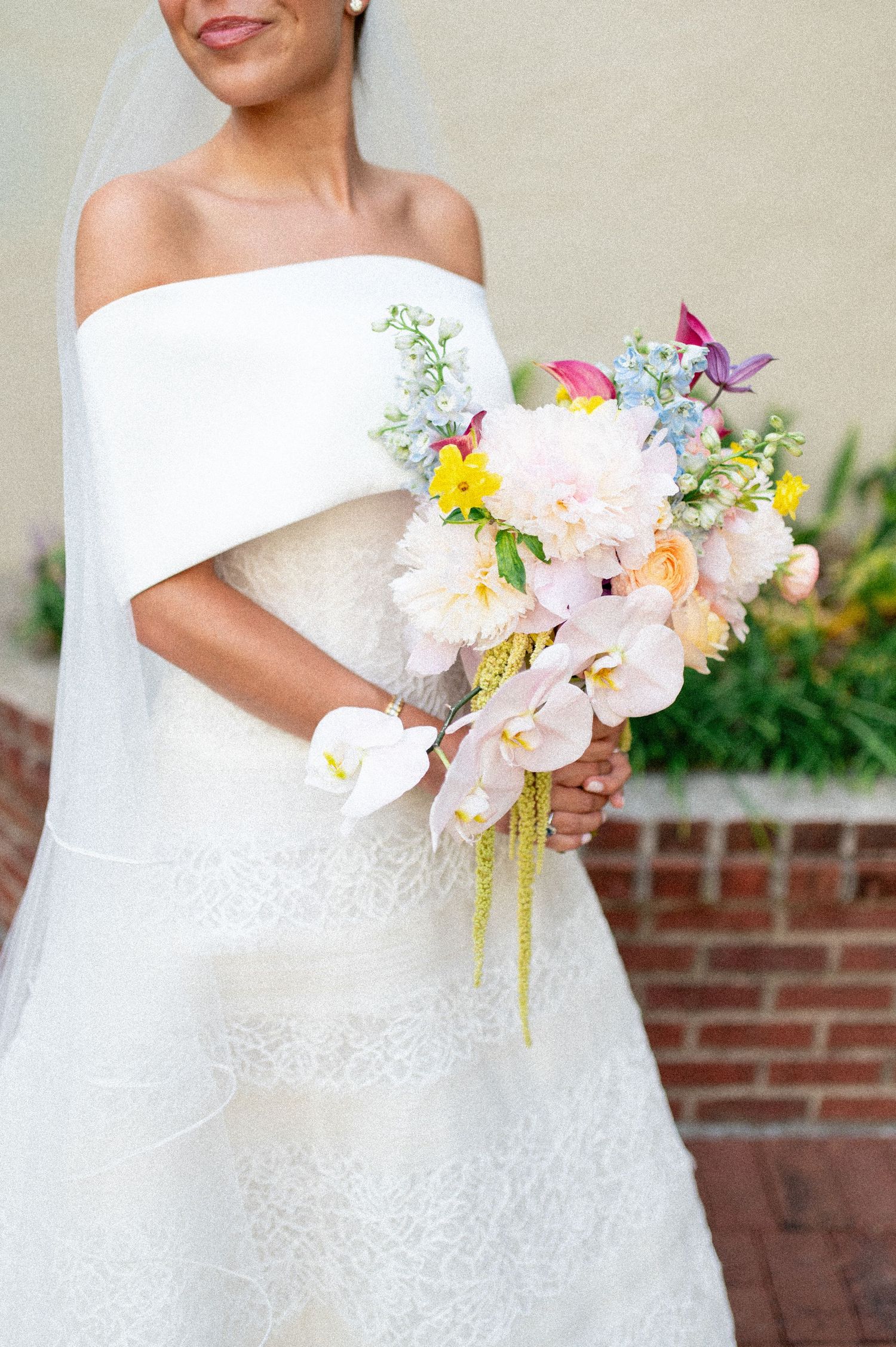Bride in off-shoulder lace wedding gown holding colorful cascading bouquet with peonies, orchids, and wildflowers.