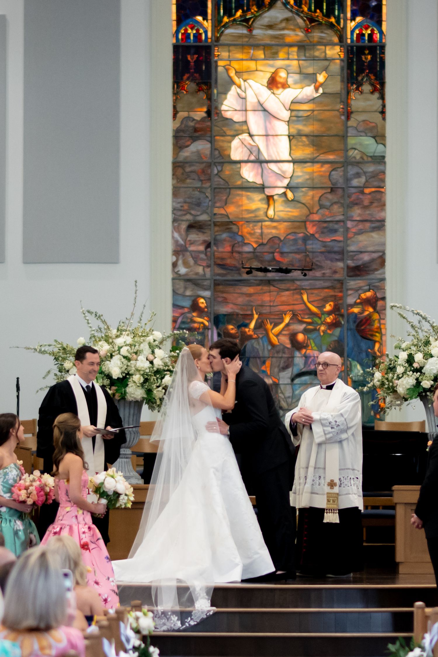 Bride and groom share first kiss at church altar before stunning stained glass Jesus window.