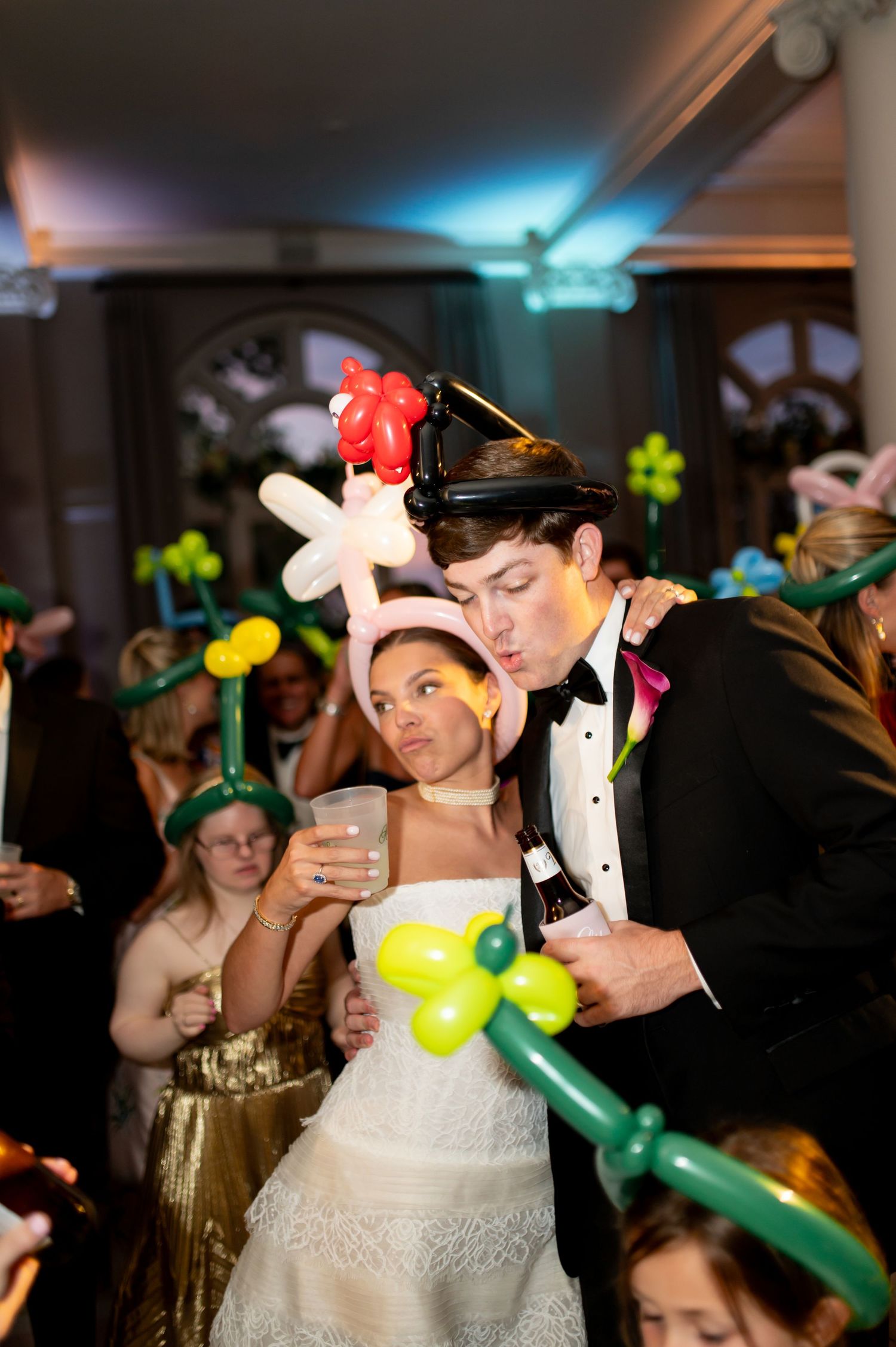 Bride and groom dancing with balloon hats amid festive wedding reception crowd at night.