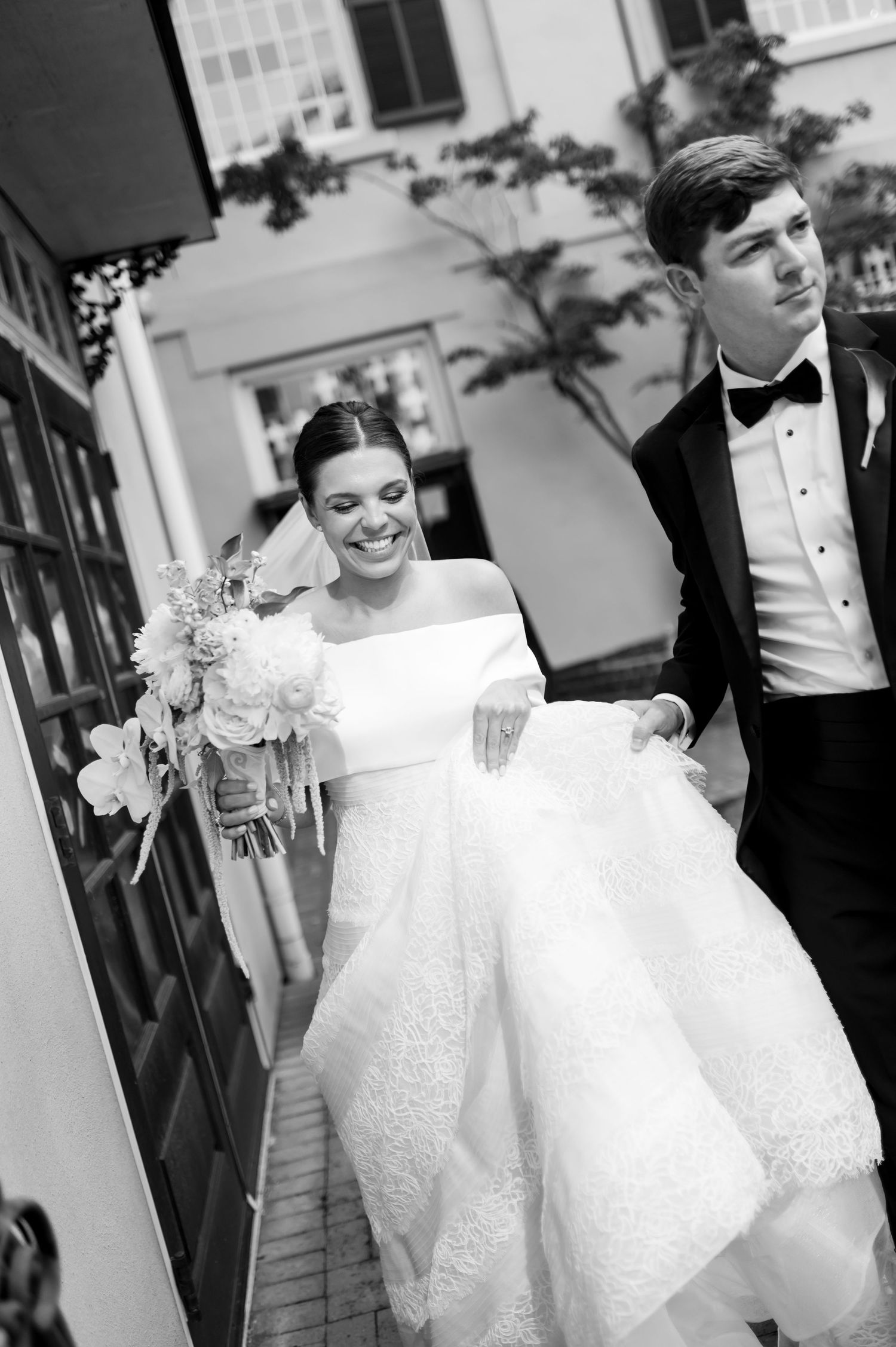 Bride and groom walk together outdoors, she holds a white bouquet in a lace gown, he wears a black tuxedo.