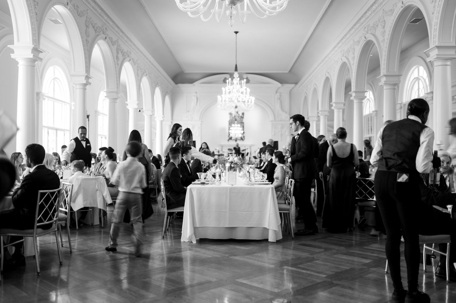 Panoramic view of an elegant white ballroom filled with wedding guests and crystal chandeliers.