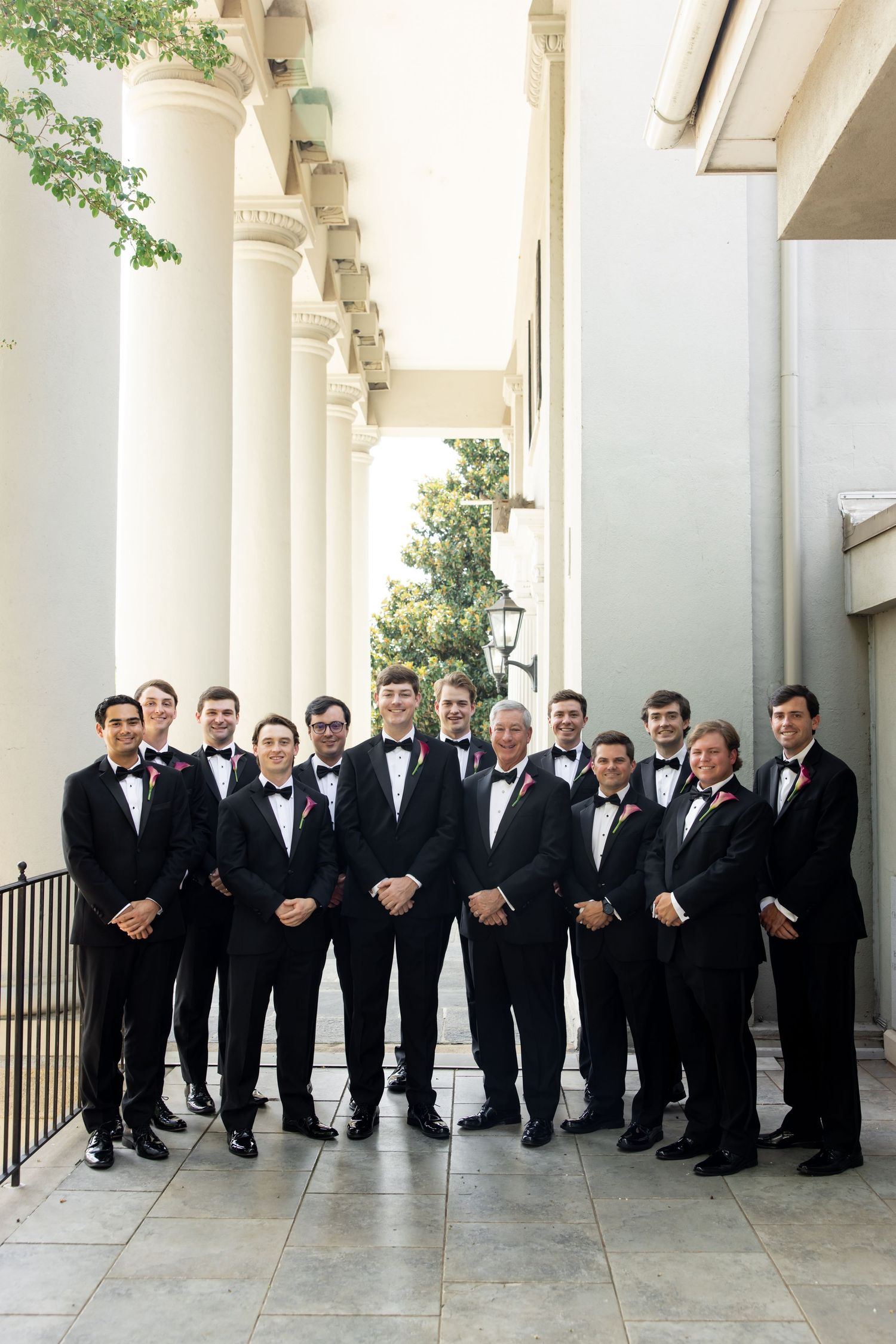 Group of groomsmen in black tuxedos with red boutonnieres posing between white columns.