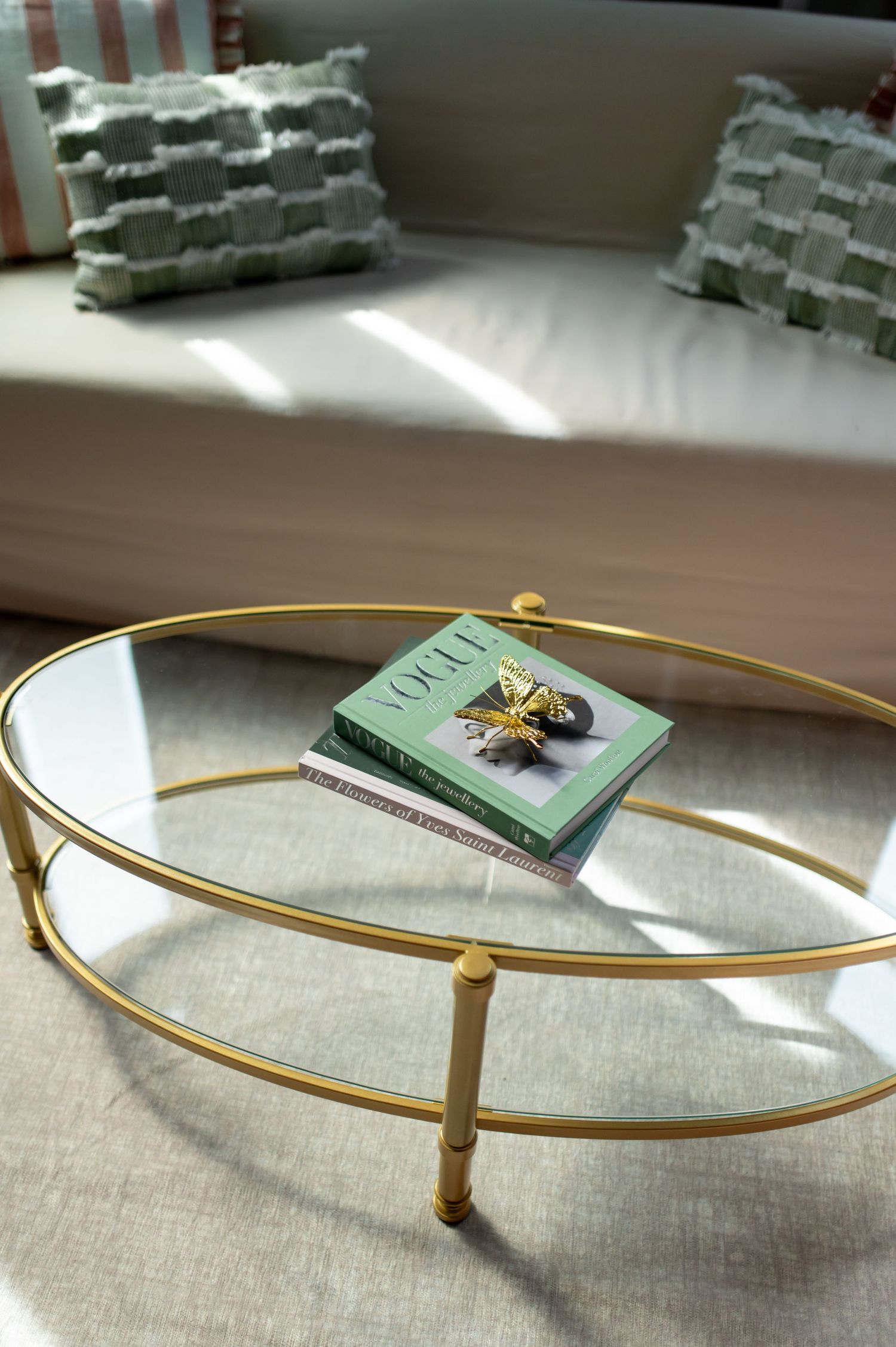 Gold oval glass coffee table with Vogue book and decorative bee figurine in a sunlit sitting room.