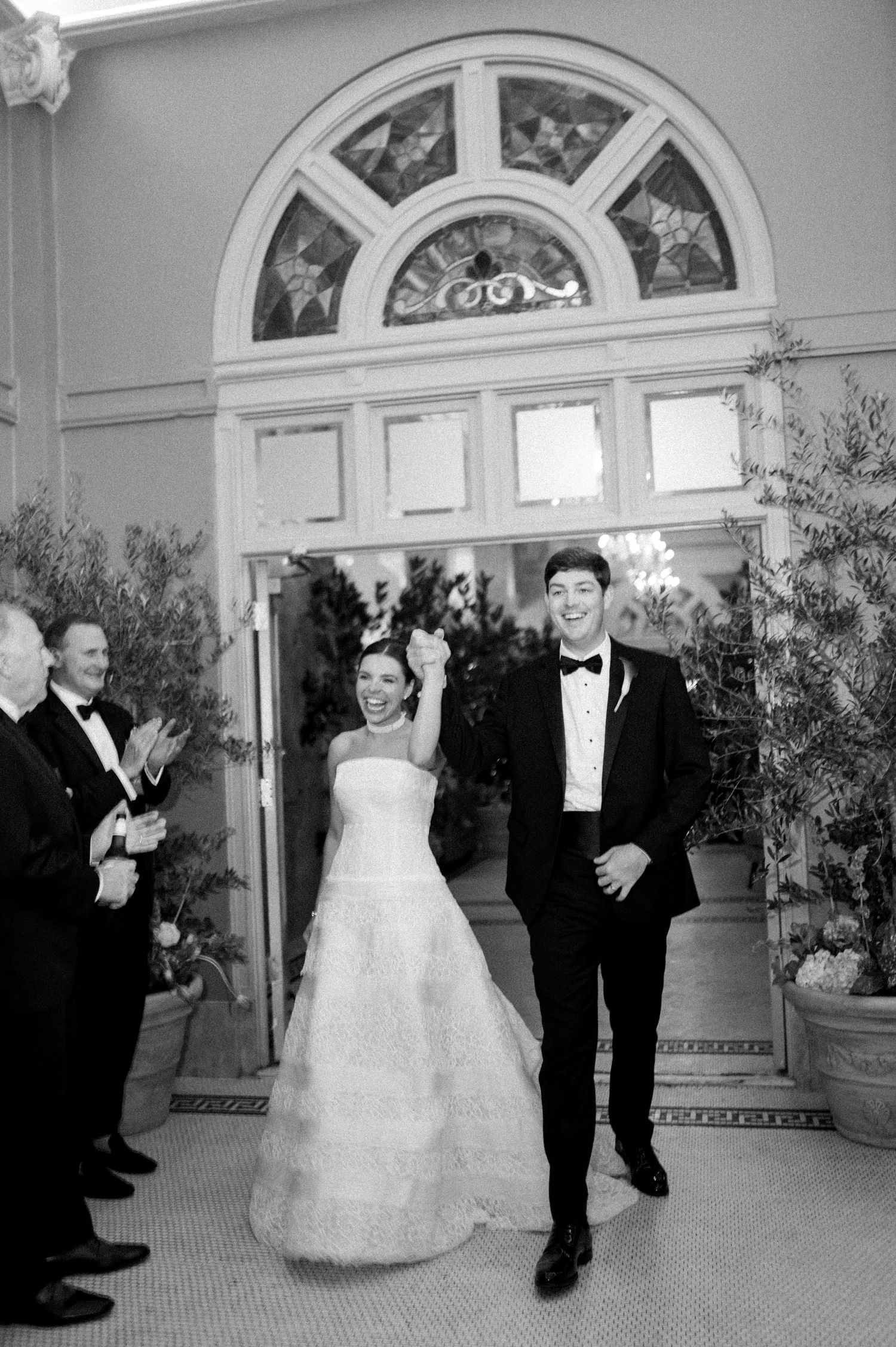 Black and white photo of jubilant newlyweds raising hands as they exit through elegant arched doorway.