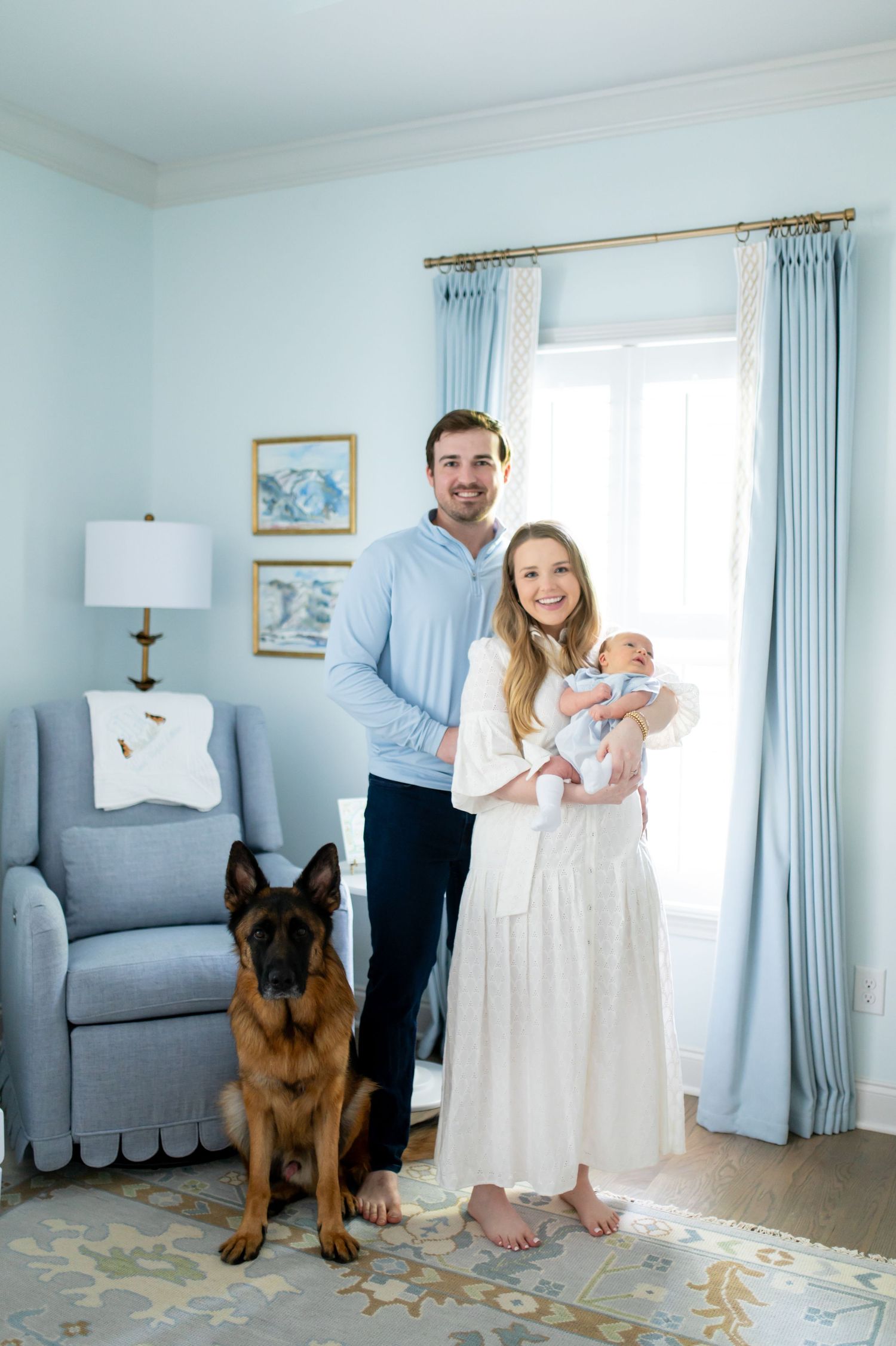 Young couple with newborn baby and German Shepherd dog posing in a light blue nursery room.