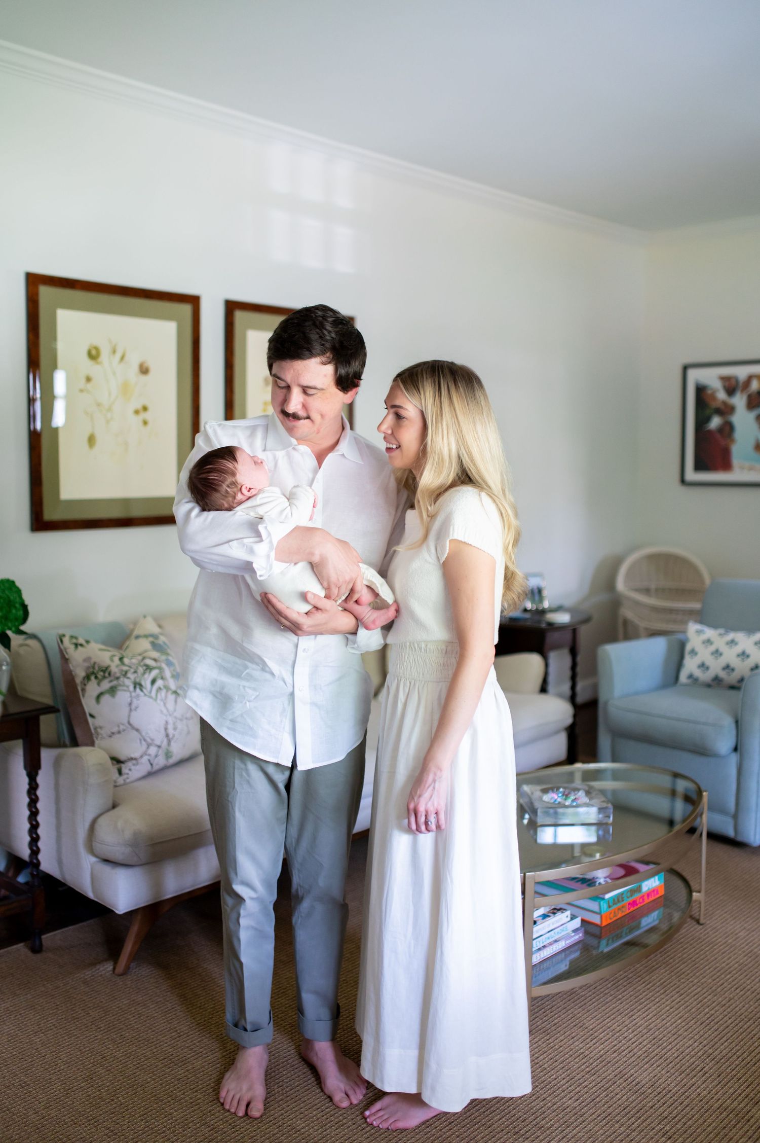 Happy couple standing indoors holding their newborn baby, smiling at each other in a bright, elegantly decorated living room.