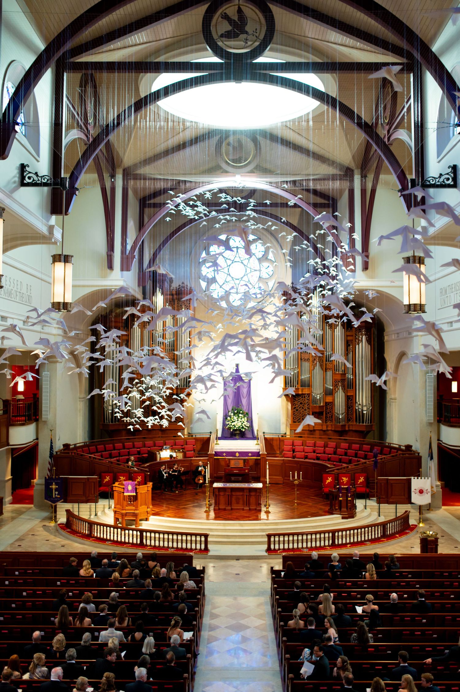 Interior of a grand church with soaring arched ceilings, rose window, pipe organ, and decorative white birds in flight above pews.