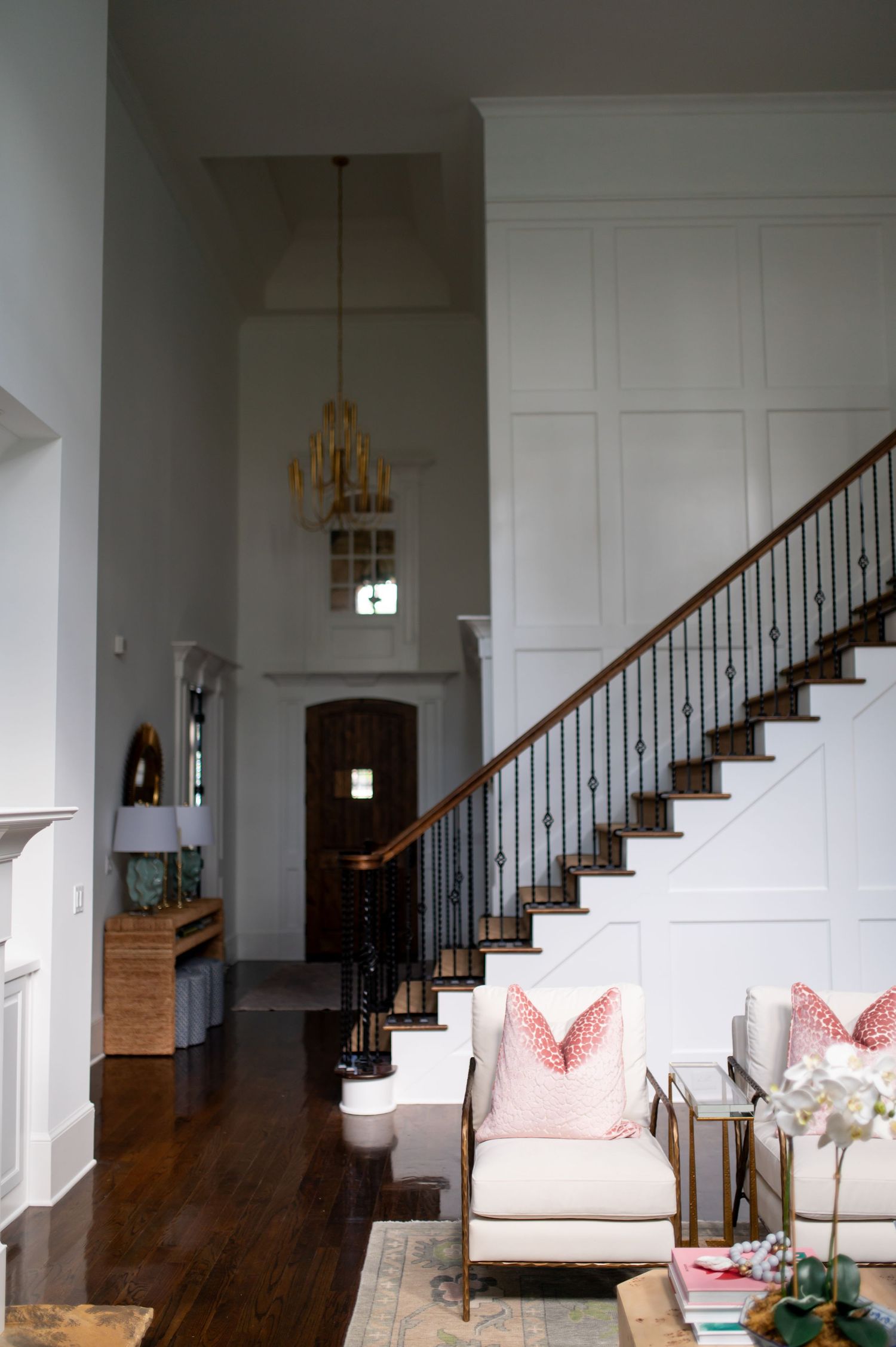 Elegant foyer with white paneled walls, hardwood floors, wrought iron staircase and a gold chandelier hanging above.