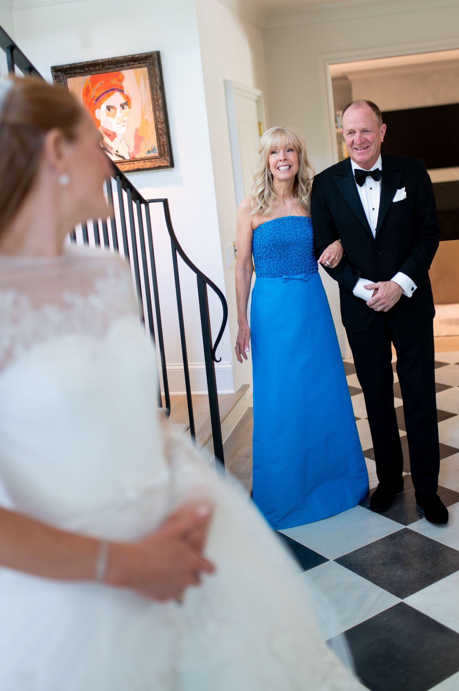 Bride reveals wedding dress to smiling parents in tuxedo and blue gown on checkered marble foyer floor.