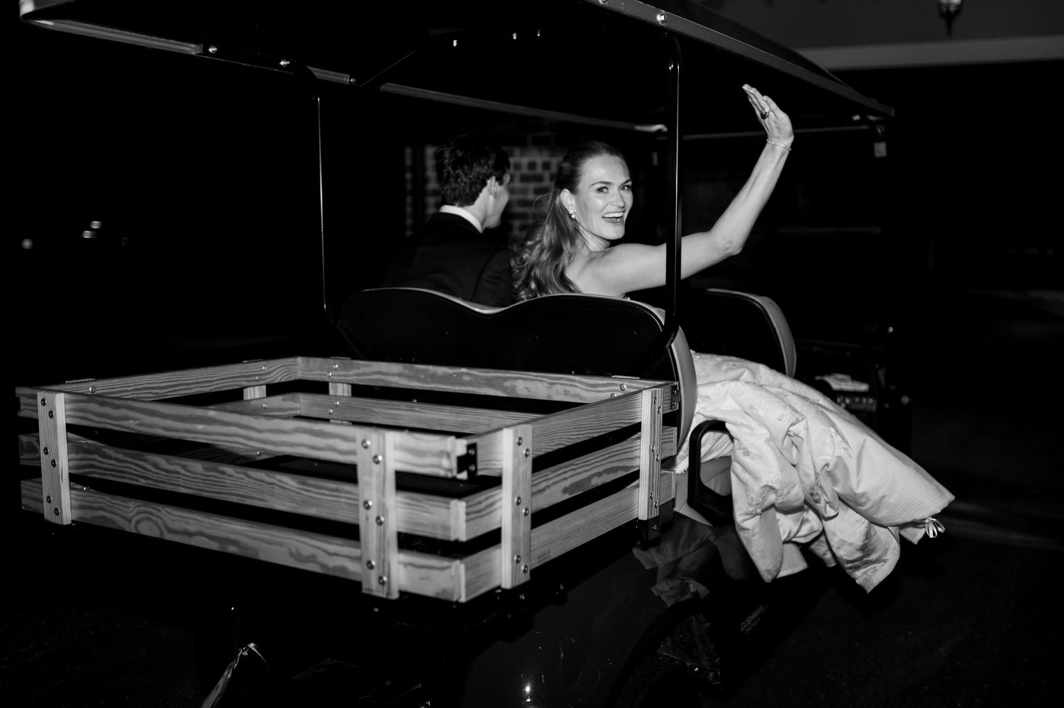 Black and white photo of a smiling bride waving from a vintage carriage at her wedding reception.