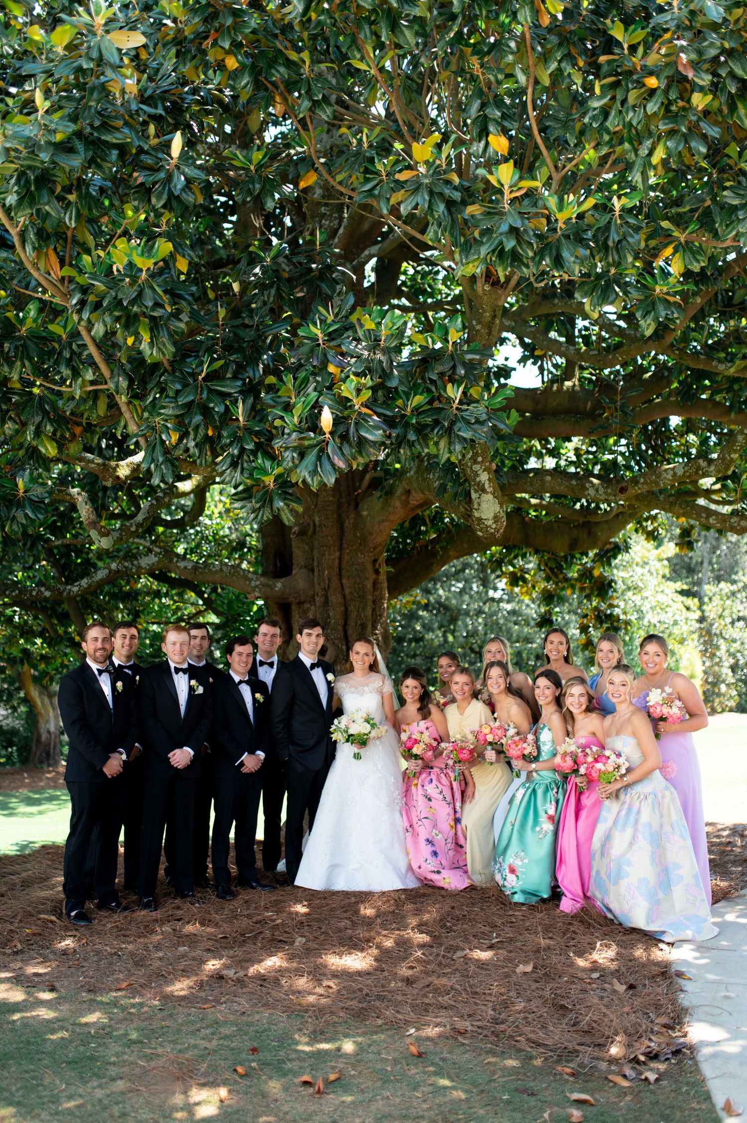 Large wedding party posing under sprawling magnolia tree with colorful bridesmaids and tuxedoed groomsmen.