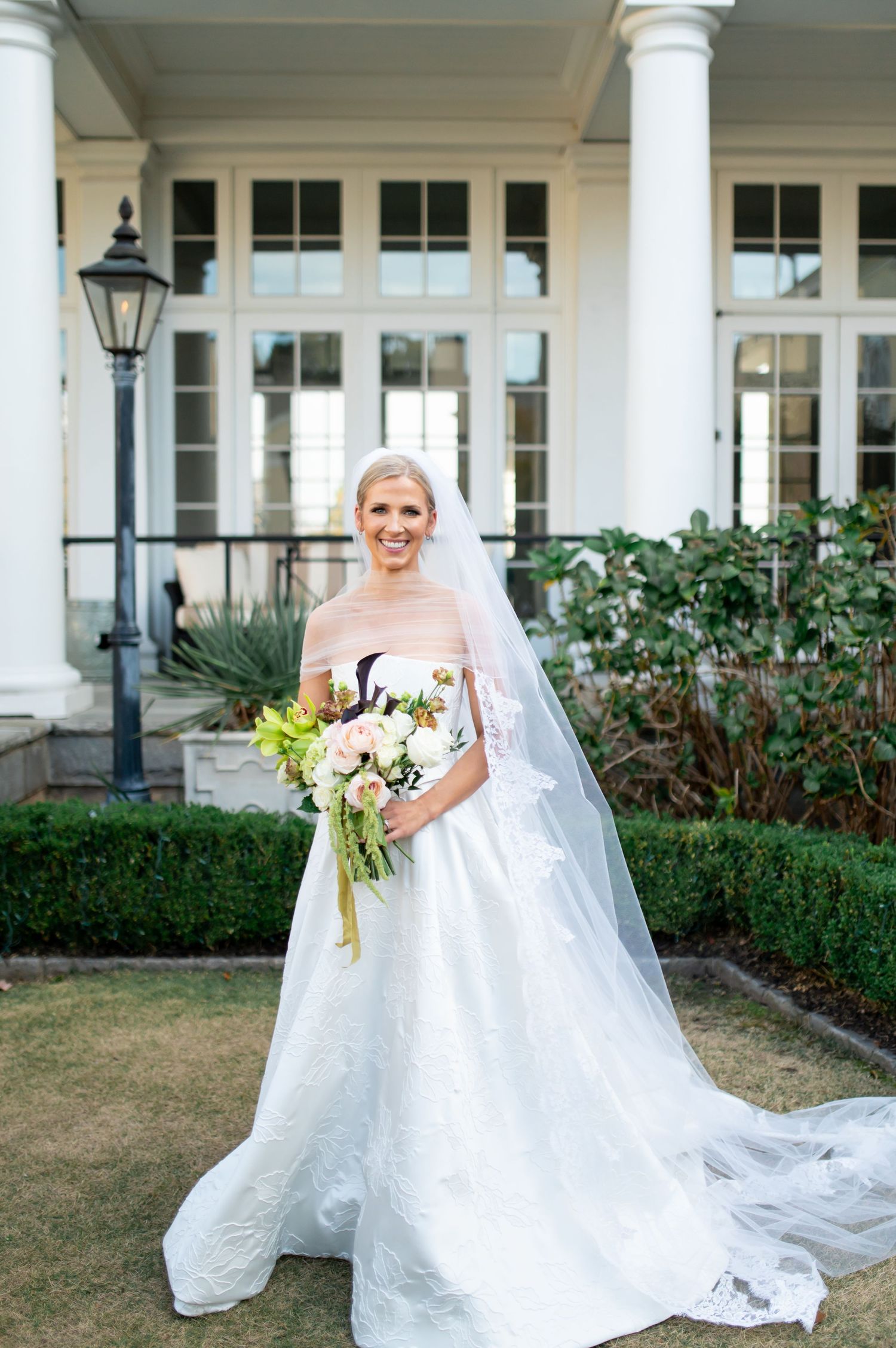 Bride in flowing white gown and veil poses in front of classic white columned building.