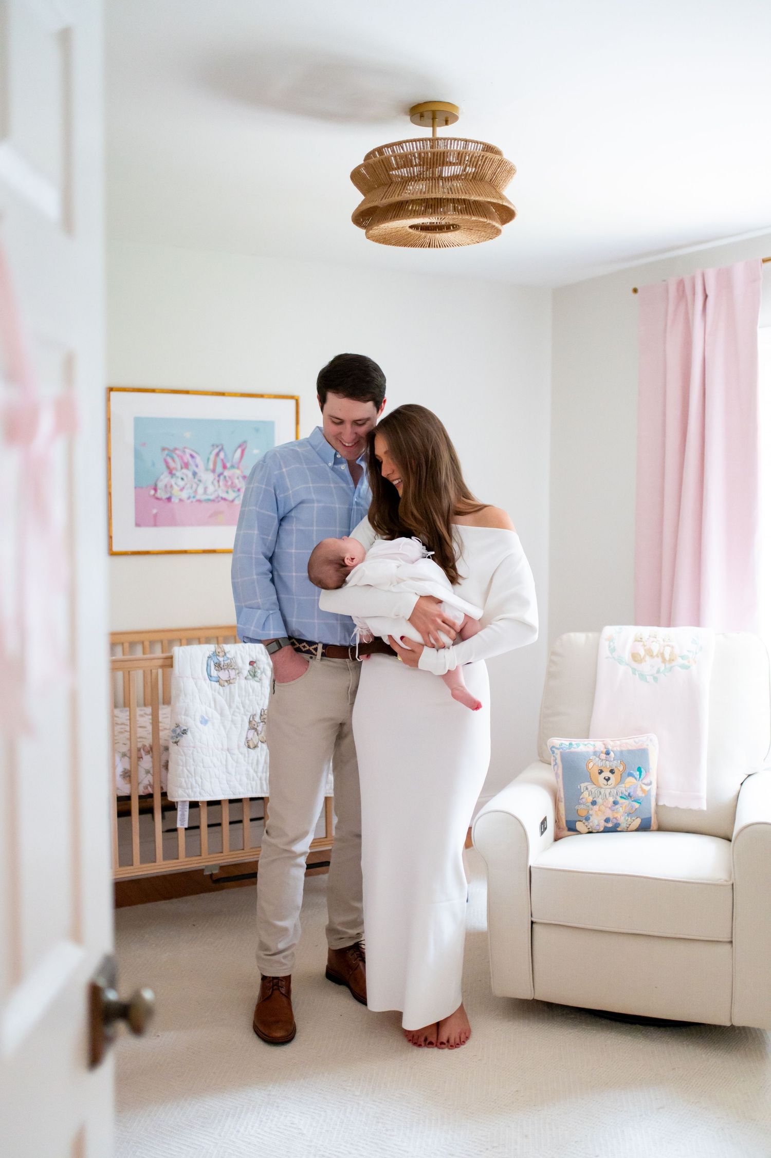 A couple shares tender moments in a bright white nursery with pink curtains and natural light streaming in.