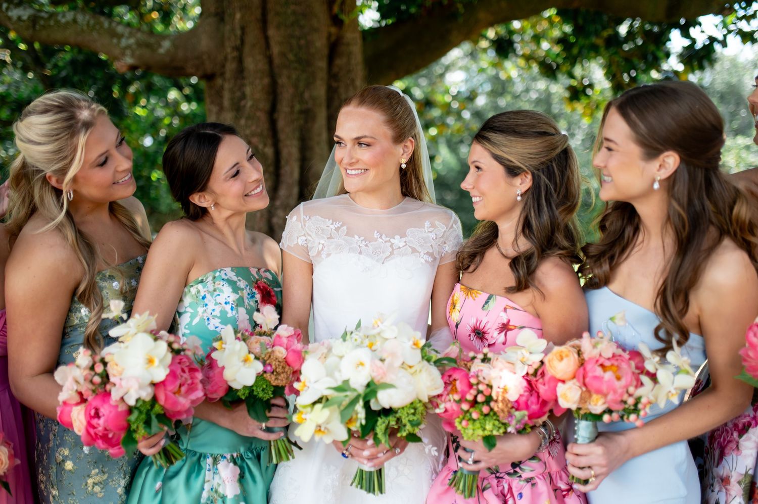Bride with bridesmaids in colorful floral dresses holding vibrant pink and white bouquets outdoors at wedding celebration.