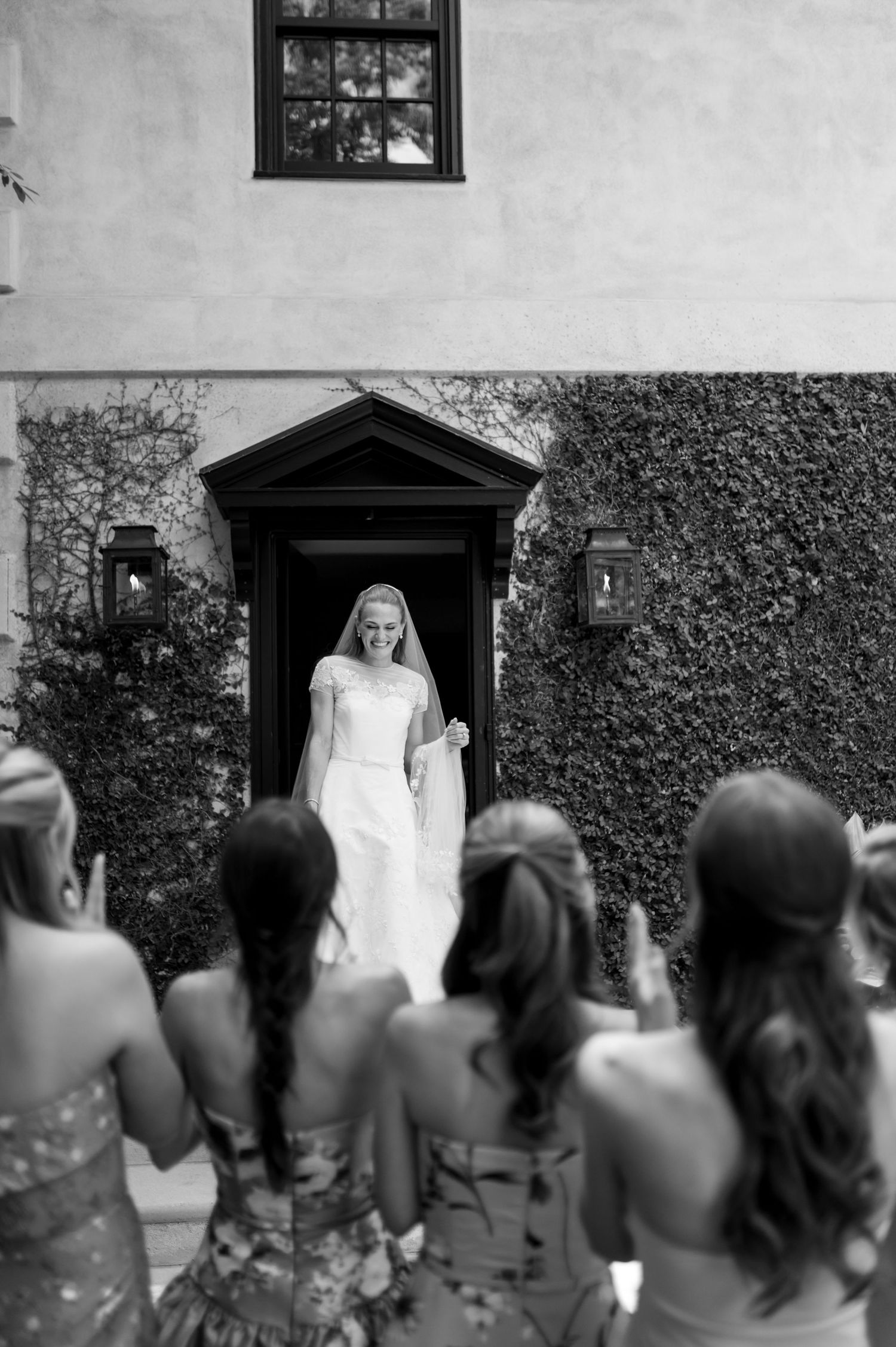 Bride in white wedding gown emerges from ivy-covered doorway as bridesmaids react with excitement, black and white photo.