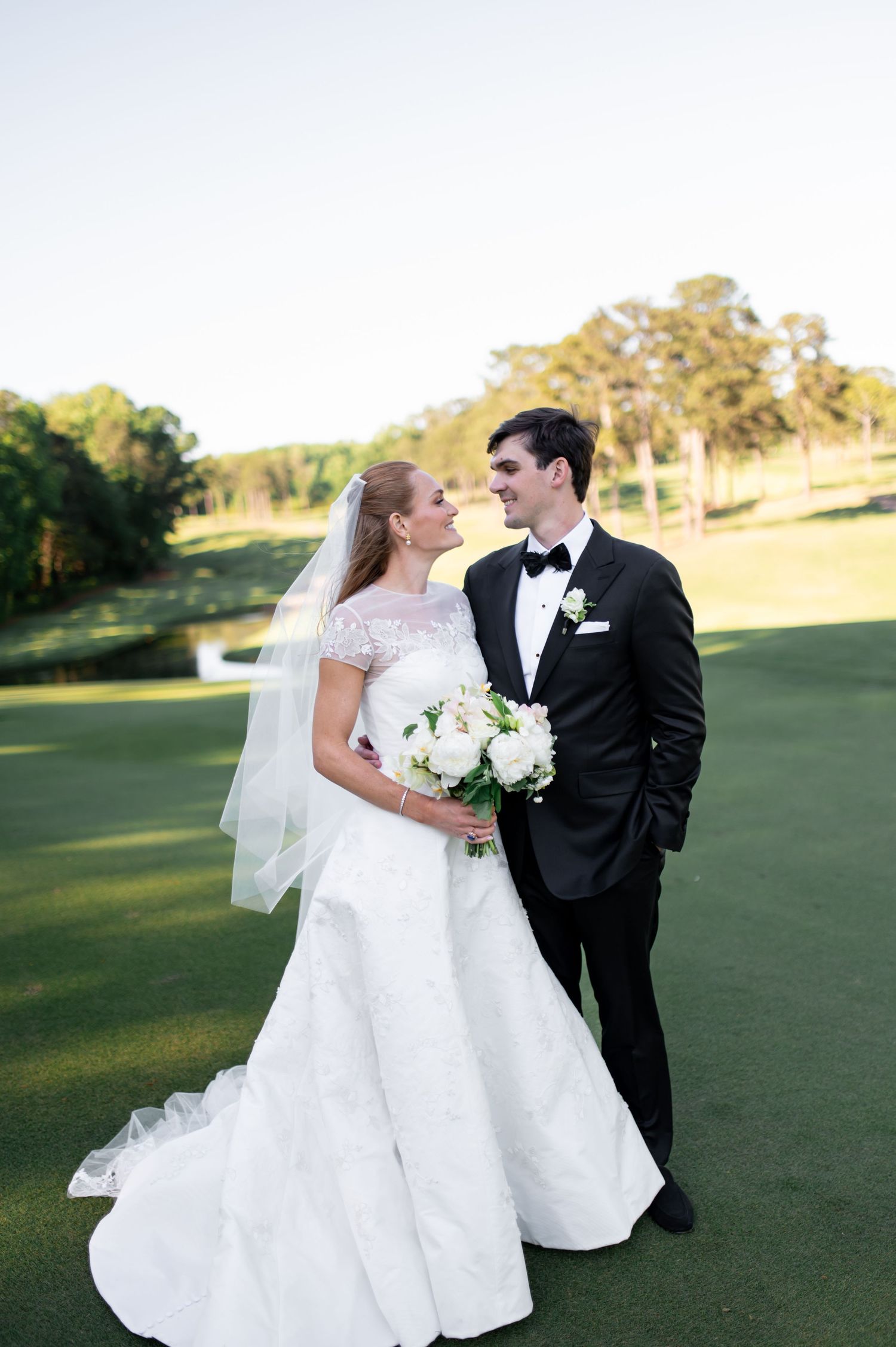 Bride and groom share romantic gaze on golf course at sunset, bride in elegant white gown with veil.