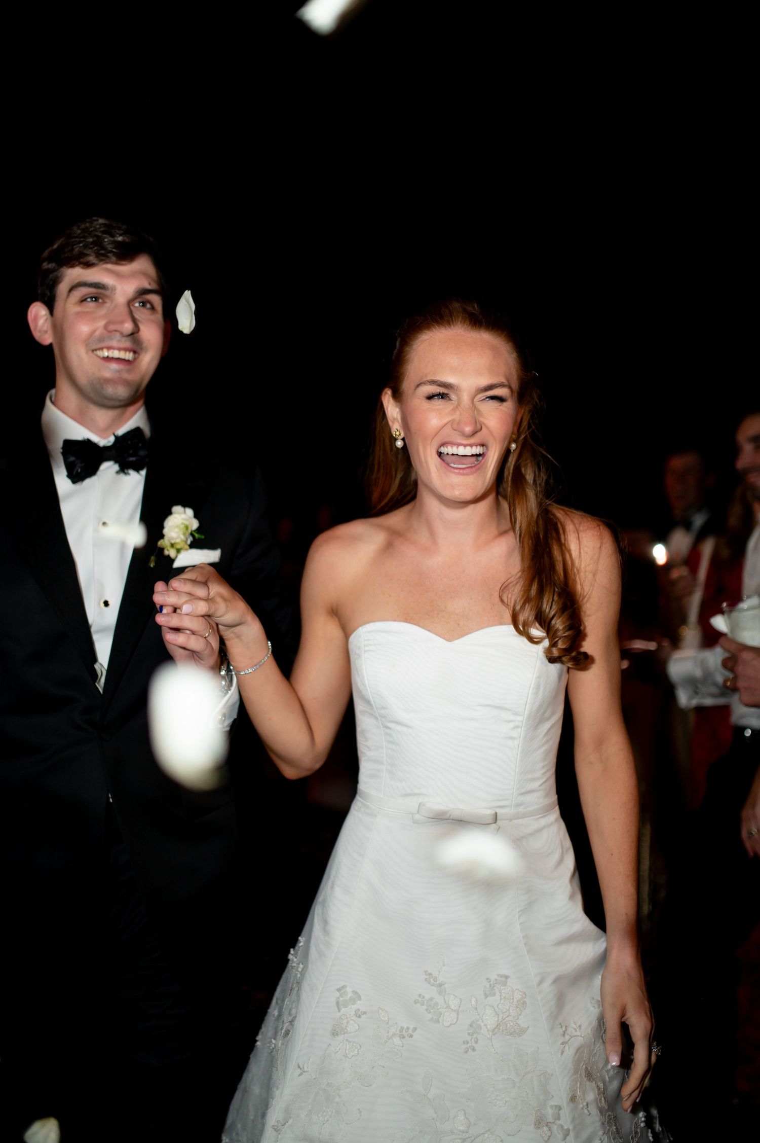Happy bride in white strapless gown and groom in black tuxedo smiling during wedding reception exit.