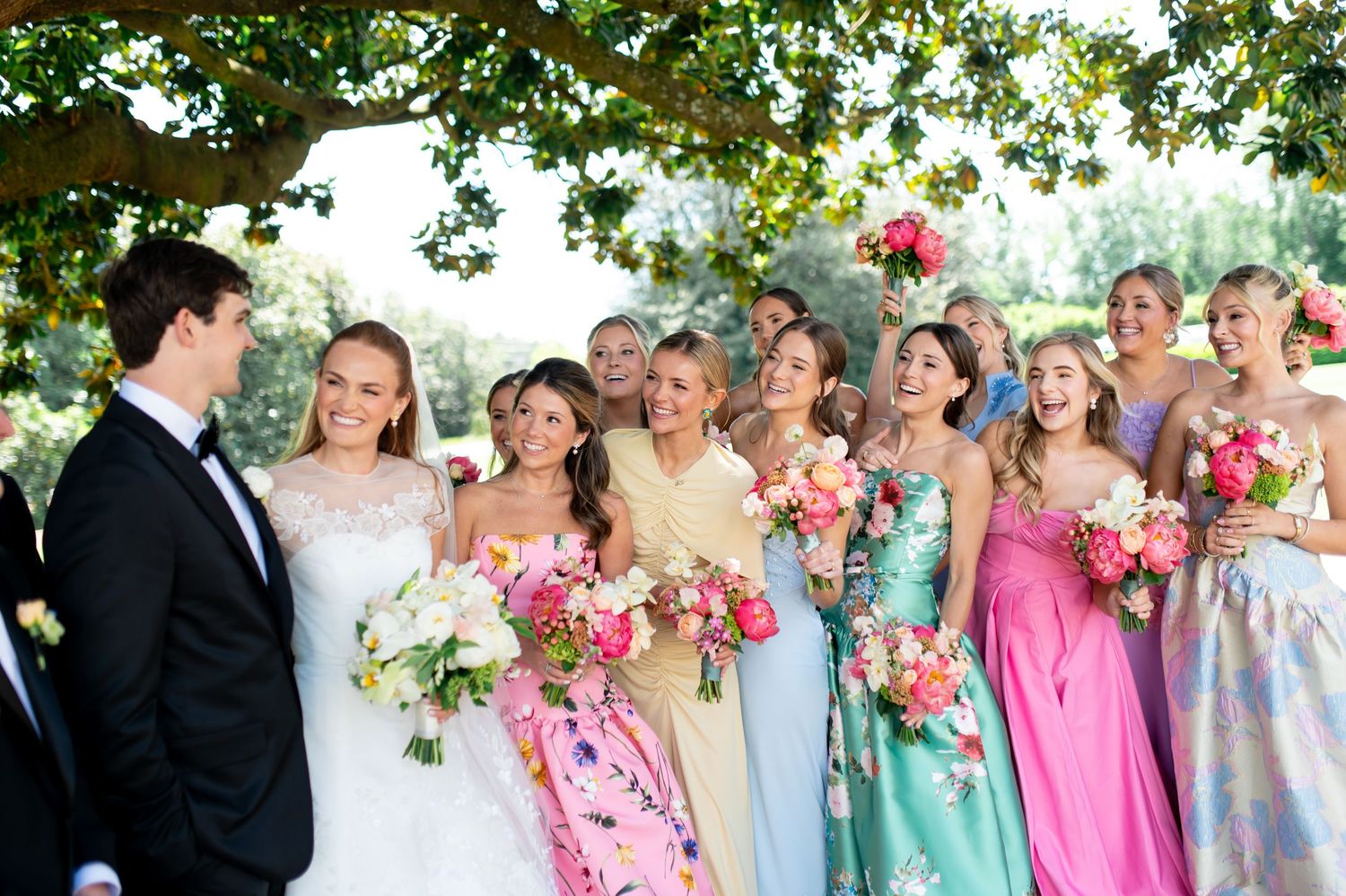 Bride and groom share joyful moment with bridesmaids in colorful floral and pastel gowns holding pink bouquets.