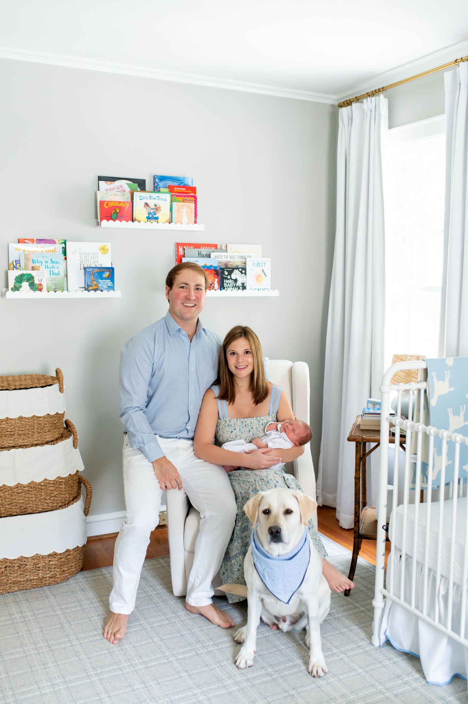 A nursery room features a white crib, bookshelves, and a couple posing with their white dog wearing a bandana.