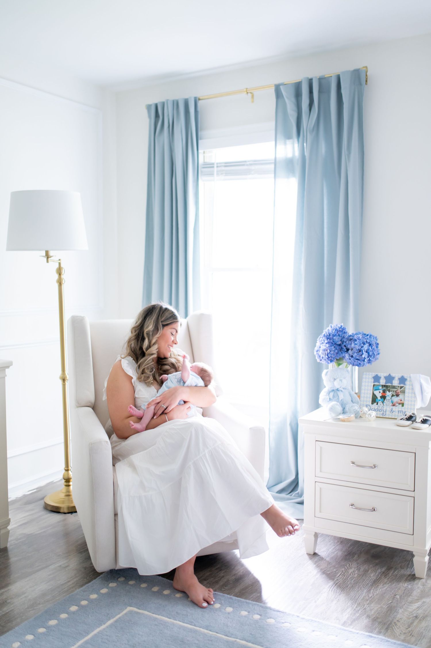 A serene nursery scene with blue curtains and someone holding a baby in a white armchair.