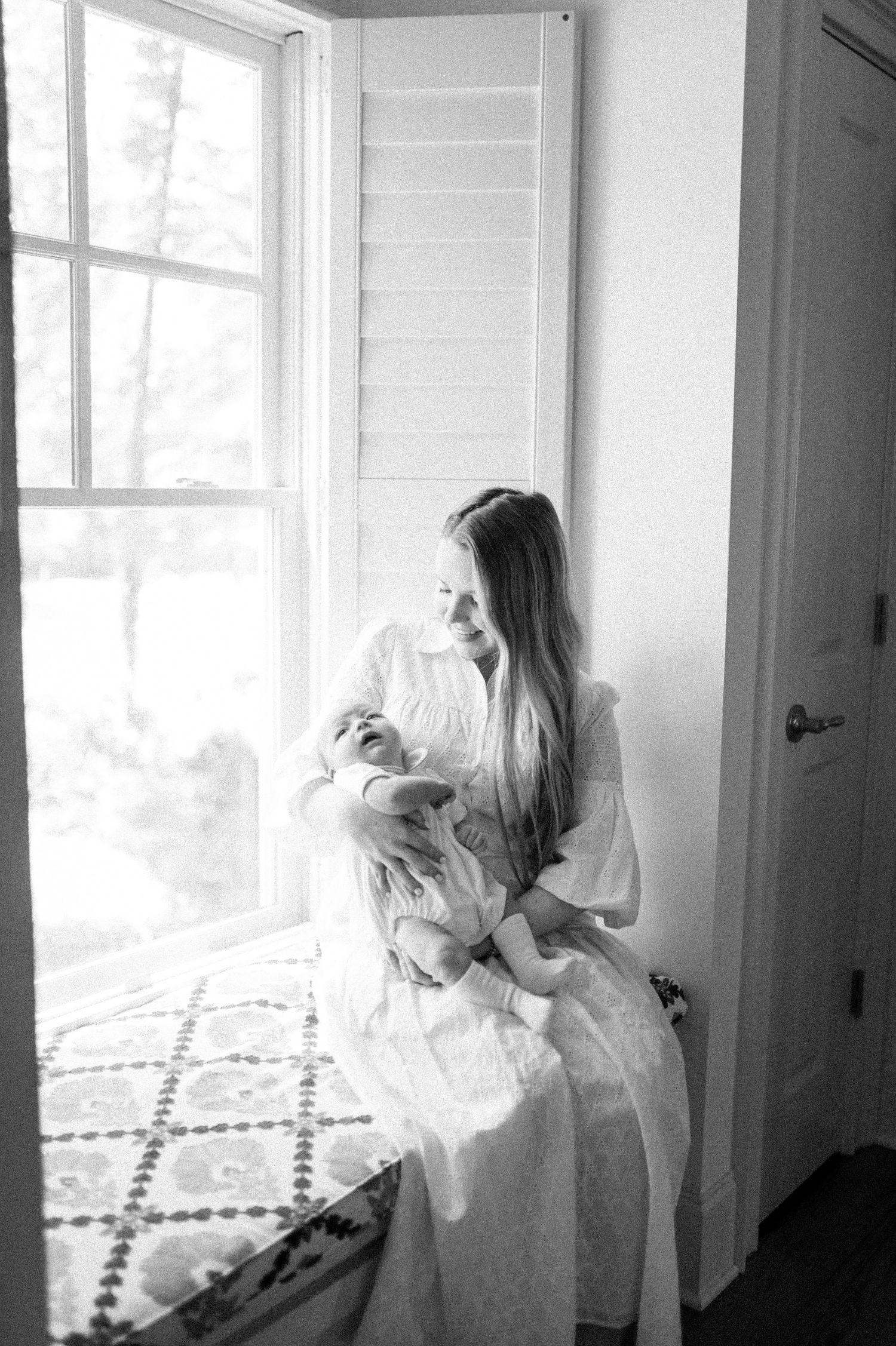 Black and white photo of a mother in white dress cradling her newborn baby by a sunlit window.