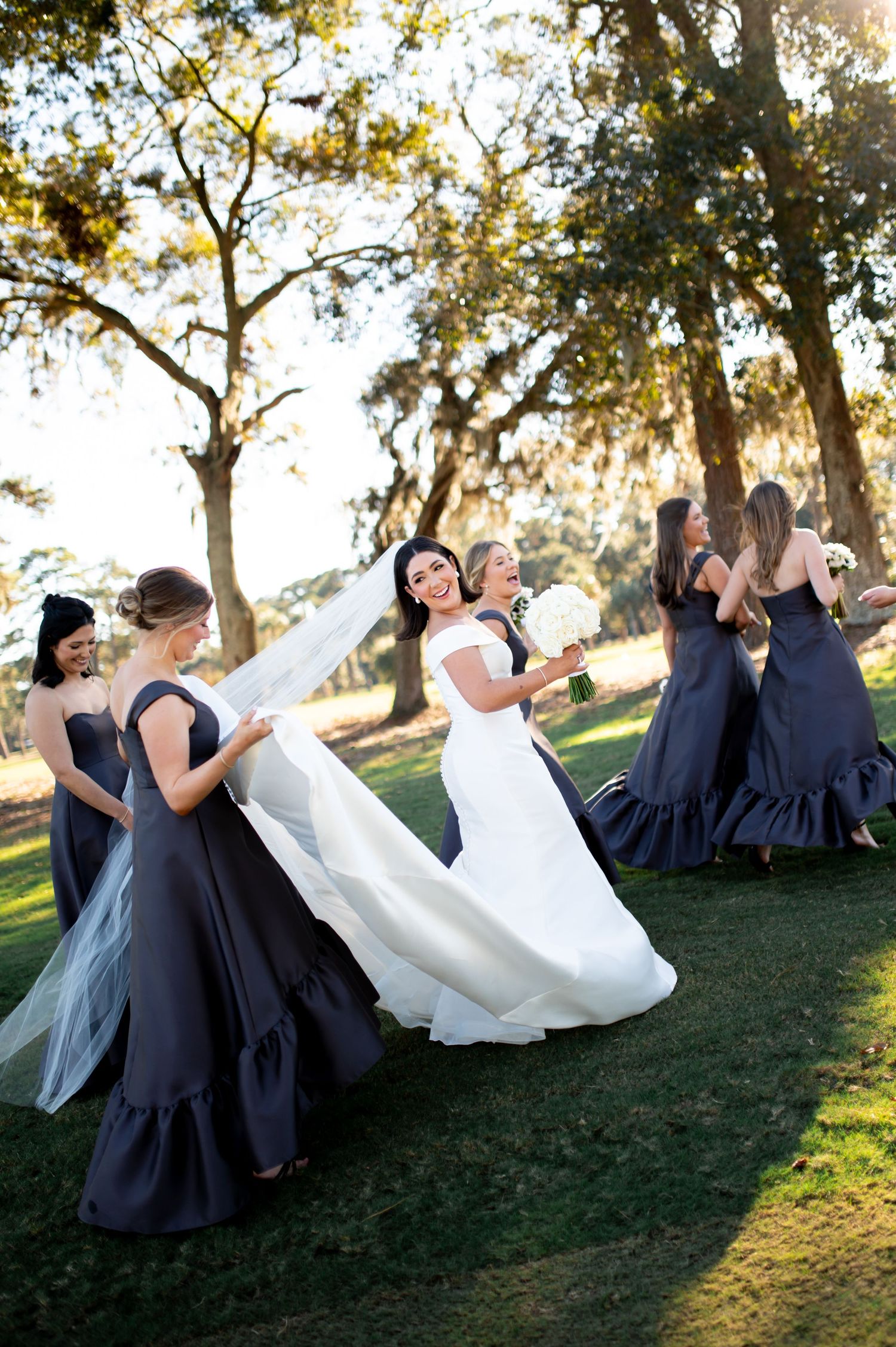 Wedding party lounges on a grassy hillside in navy dresses and a white gown during sunset.