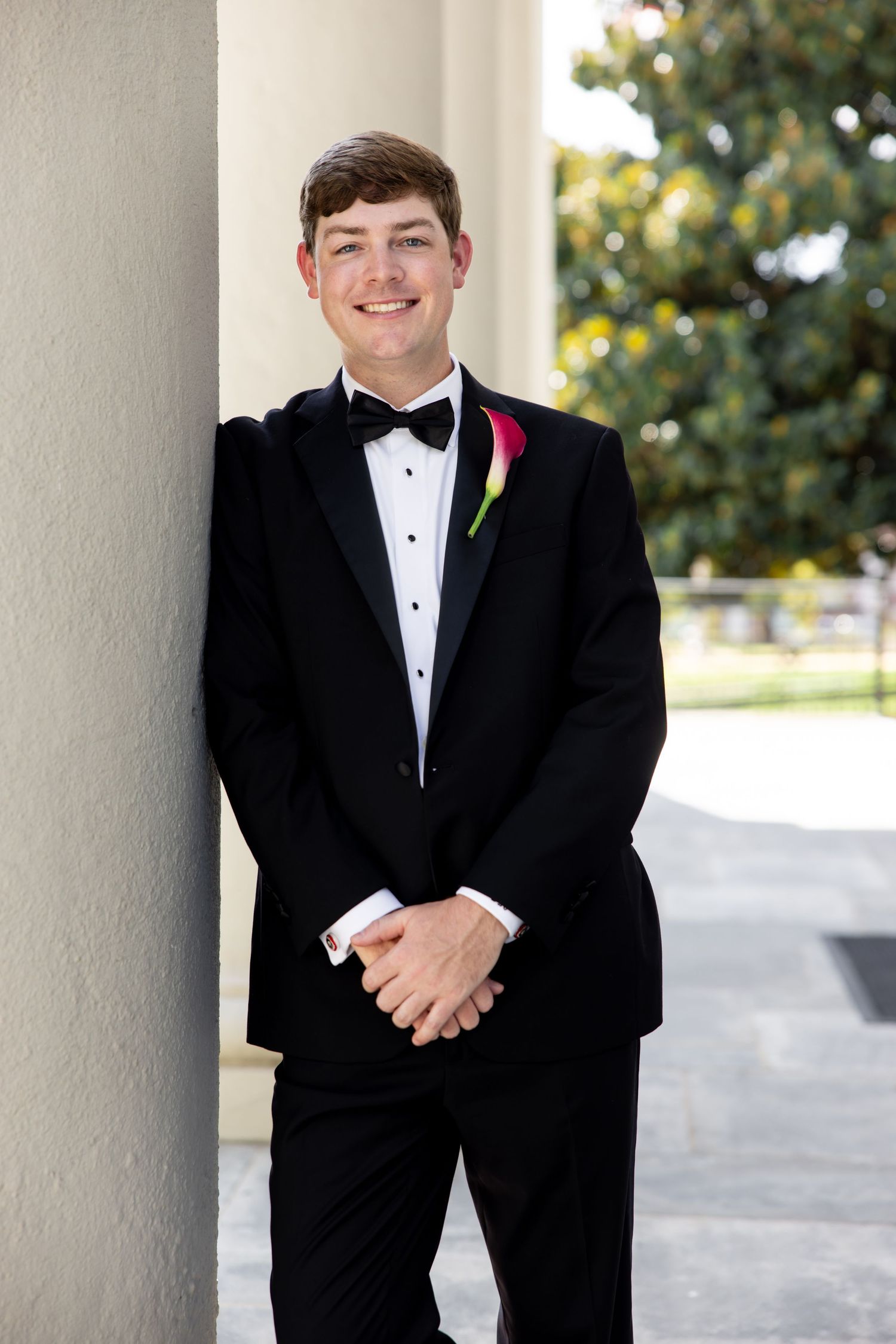 Young groom in black tuxedo with bow tie and rose boutonniere leaning against white column outdoors.