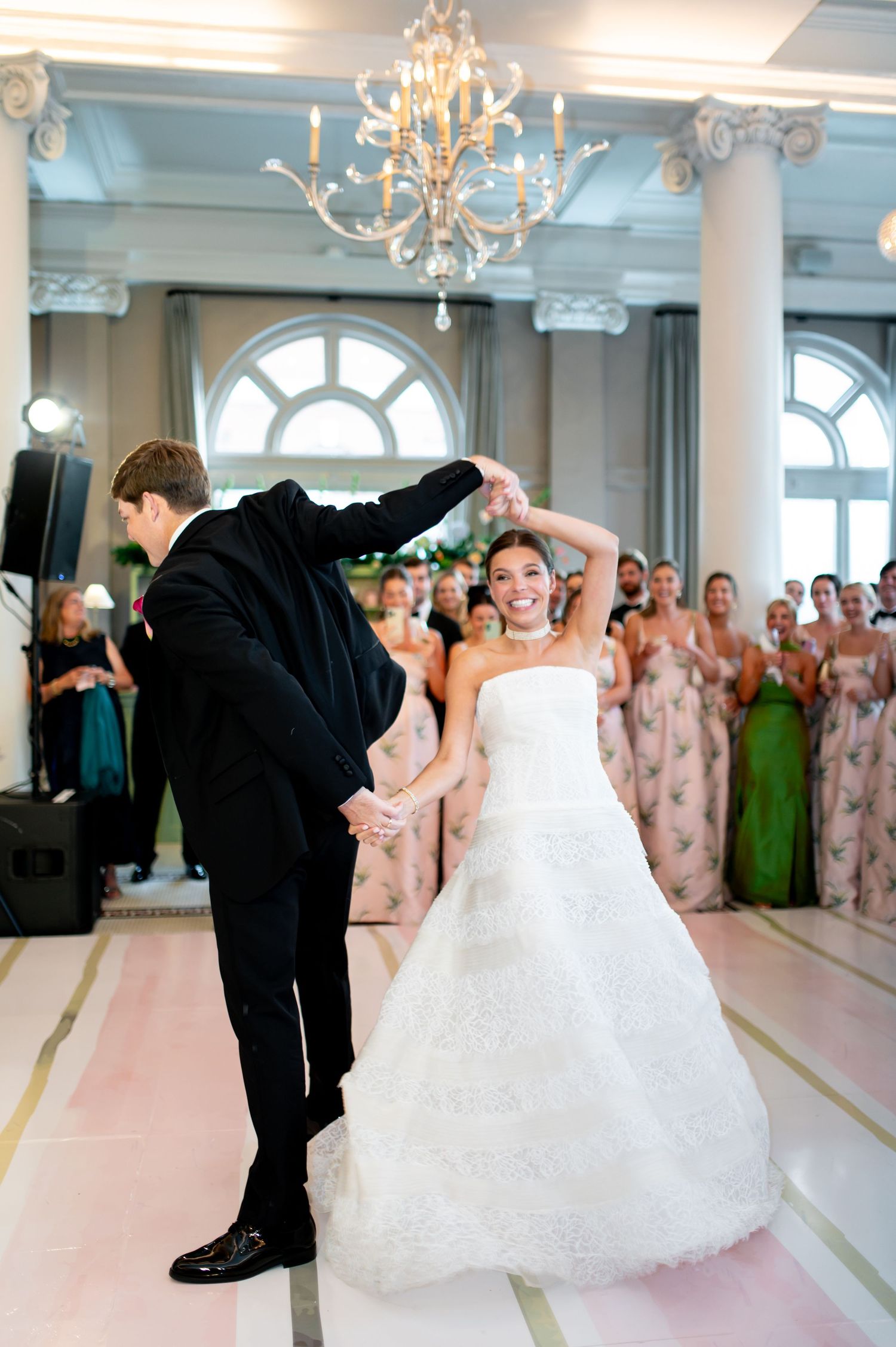 Groom twirls bride in white lace gown during first dance under chandelier with guests watching.