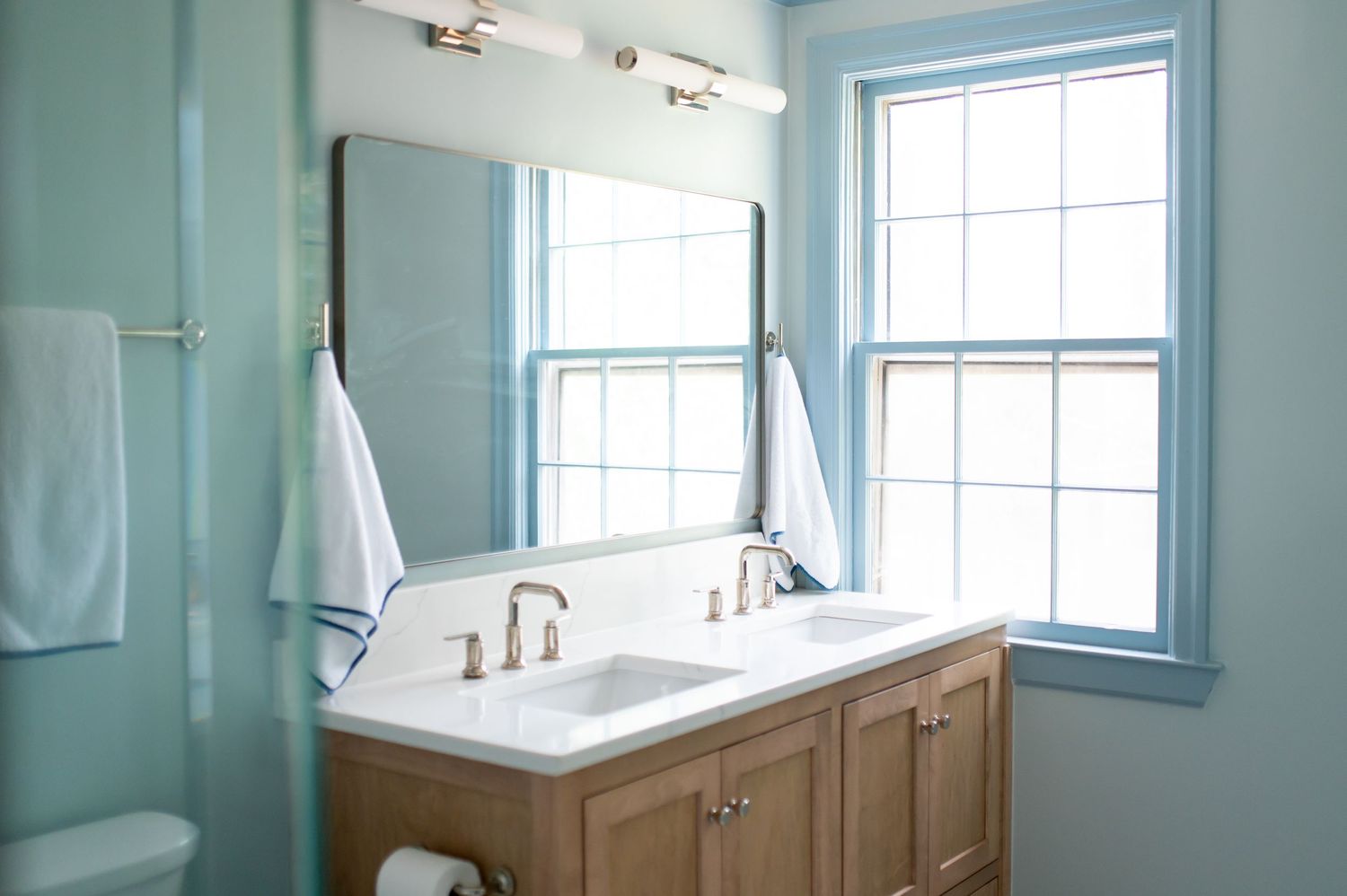 A modern bathroom with light blue walls features a freestanding tub, white fixtures, and multiple windows letting in natural light.