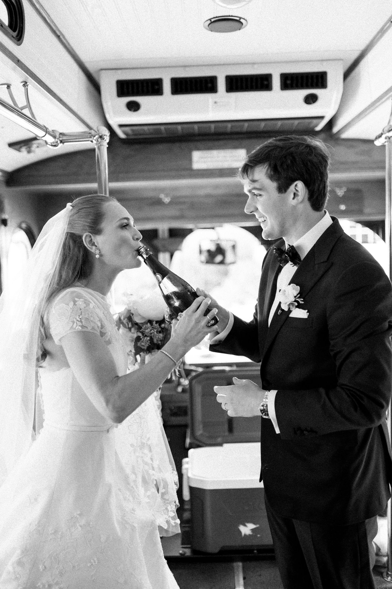 Bride and groom share champagne bottle drink on vintage bus in black and white wedding photo.