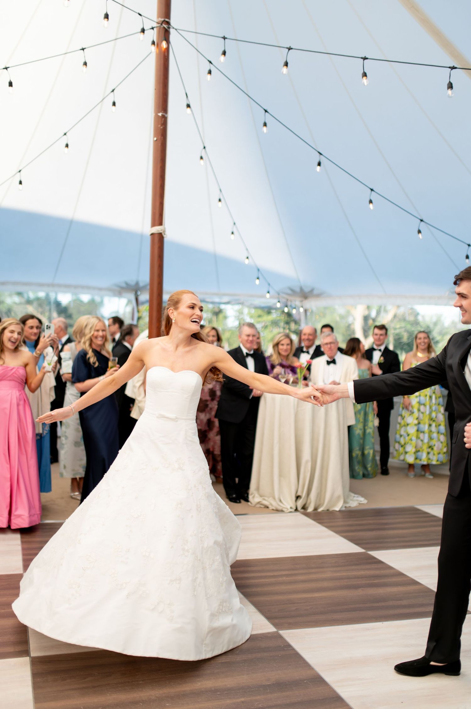 Bride in white ball gown twirling joyfully during first dance under string lights in wedding tent.