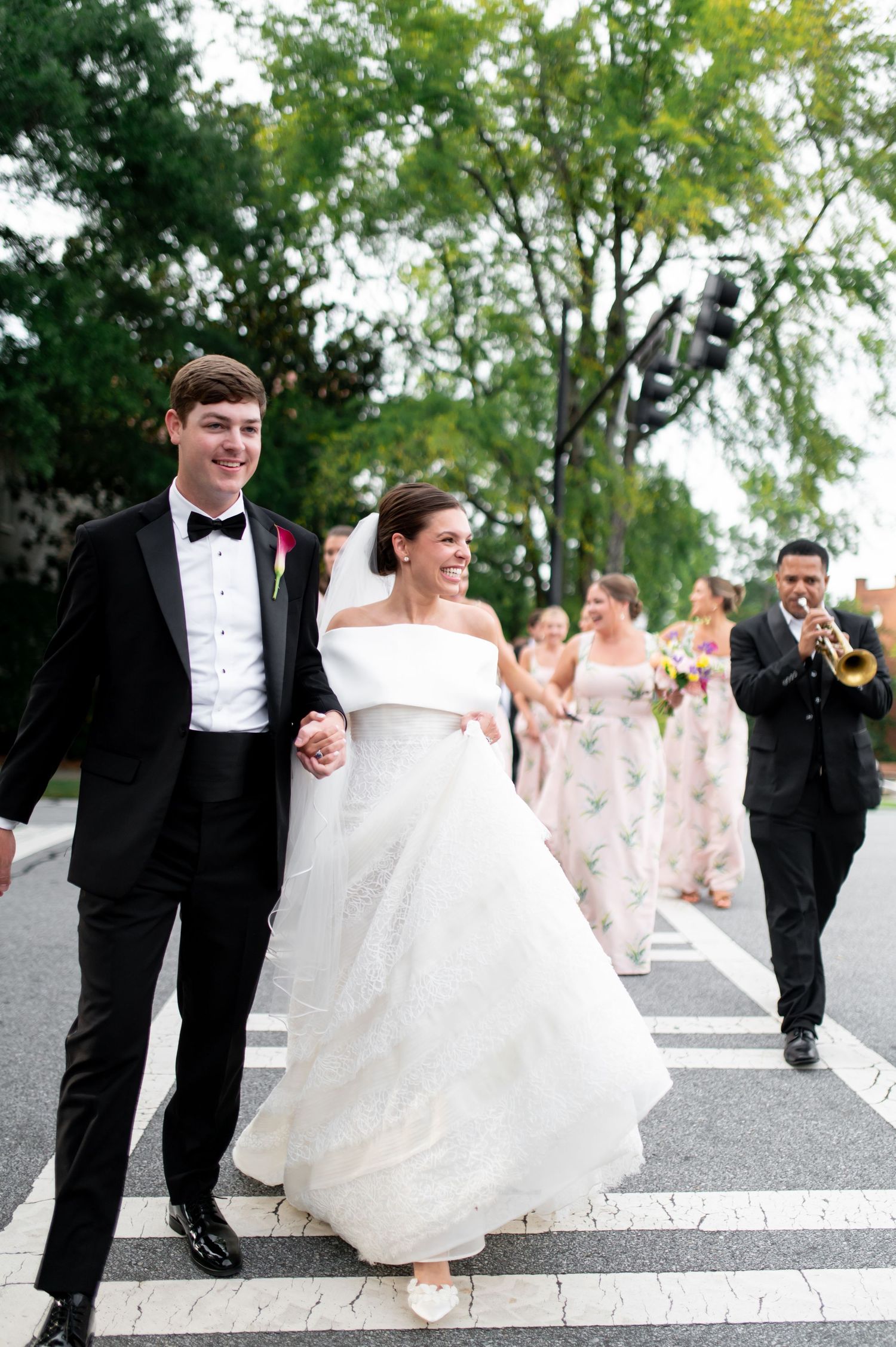 Smiling newlyweds walk hand-in-hand across street with bridal party and trumpet player following.