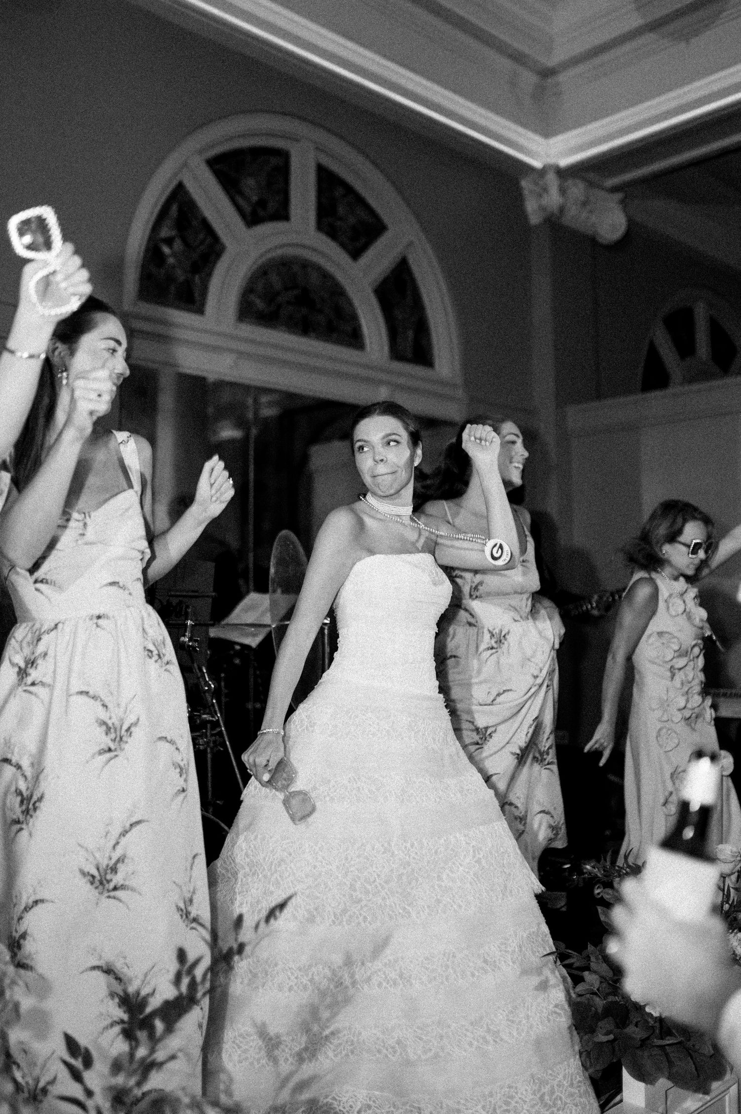 Black and white photo of bride dancing joyfully with bridesmaids in floral gowns at reception.