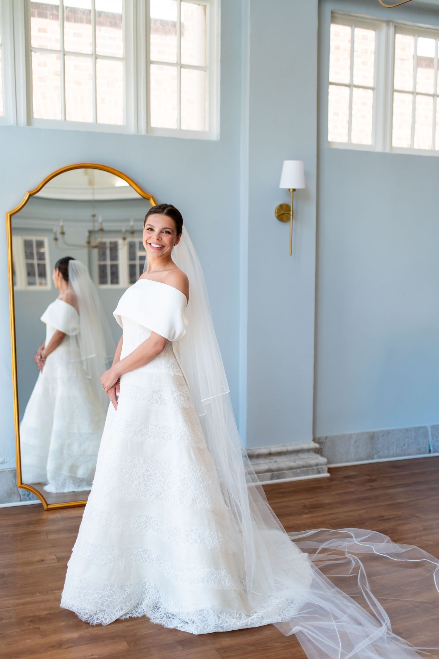 Smiling bride in off-shoulder gown and long veil stands before gold mirror in blue bridal suite.